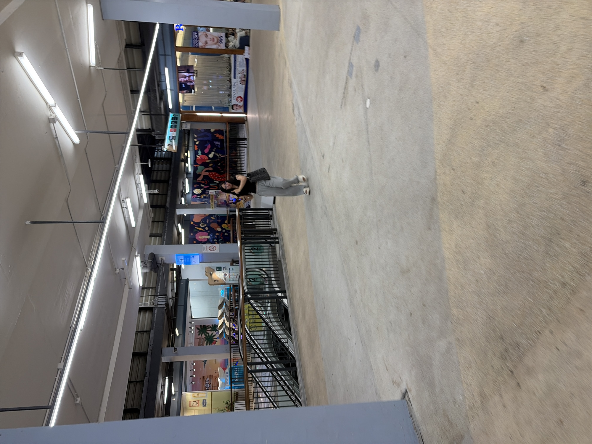 The image shows a person standing in a well-lit indoor space, likely a mall or market, in Baan Chaweng, Thailand. The person, wearing a black top and grey pants, is holding a dark bag and appears to be looking down or away from the camera. The setting is a wide walkway with a textured concrete floor. On the left side of the walkway, there are shops with colorful displays and signage. One sign has an image of a woman and the text "50% OFF". Further down, there's a graphic display with abstract shapes and figures, and another sign with images of palm trees. On the right side, the walkway continues, and a black metal railing indicates a lower level or an escalator. The lighting suggests it's daytime, and the indoor setting implies no specific weather conditions are visible. The overall mood is casual and everyday.