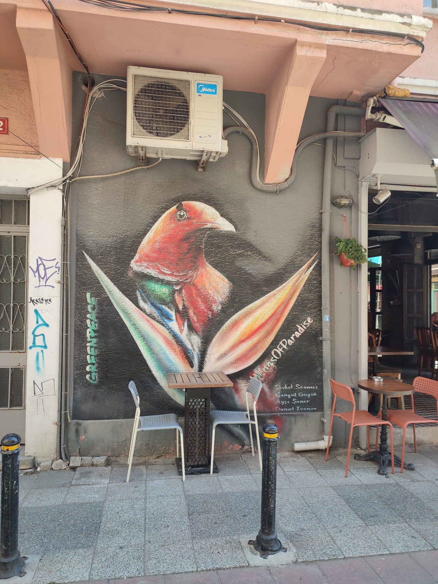 The image shows a mural of a bird on a dark-colored wall in Kadıköy, Turkey. The bird has red and orange plumage and appears to be a stylized representation of a bird of paradise. The mural is located on the side of a building that has an air conditioning unit mounted above it. In front of the mural, there are two tables and four chairs, suggesting an outdoor seating area for a cafe or restaurant. The tables are made of wood and metal, and the chairs are modern and minimalist in design. There are also several bollards along the sidewalk. The overall impression is one of an urban environment with artistic elements.