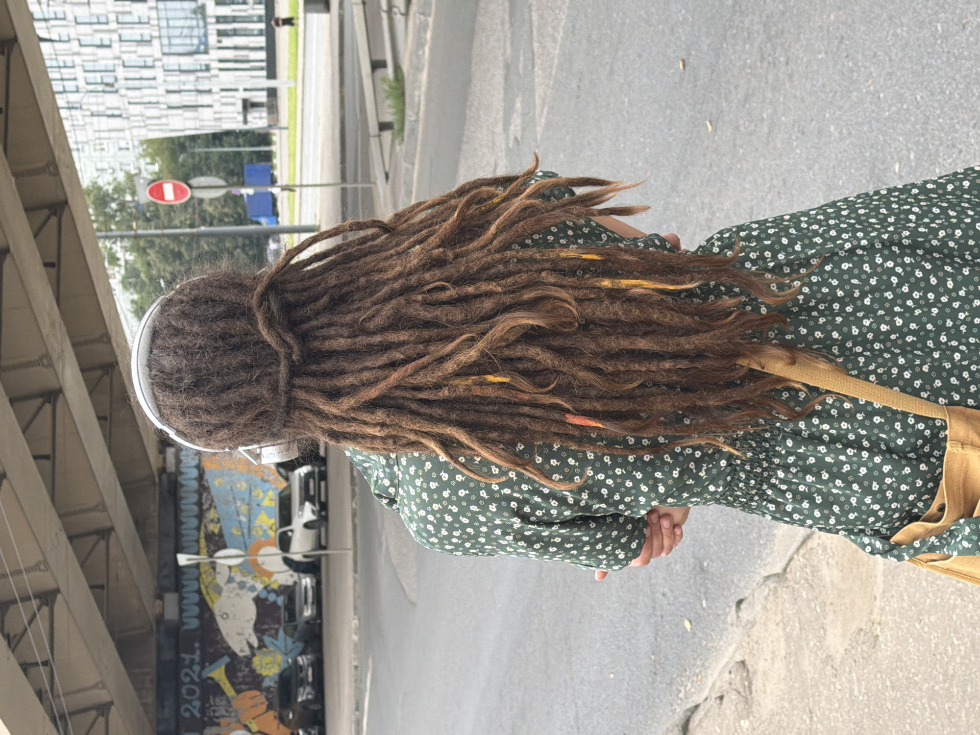 A person with long, brown dreadlocks, wearing a green floral dress and white headphones, stands facing away from the camera on a city street. The background includes a concrete overpass with graffiti, several parked cars, a modern building with many windows, trees, and street signs. A beige crossbody bag is visible on the person's right side. The street appears to be an intersection or a busy road, with a clear day sky overhead. The graffiti on the overpass includes "2021." and various artistic images. A red and white "No Entry" sign is visible on a pole in the background. The person is standing on a sidewalk next to the road.