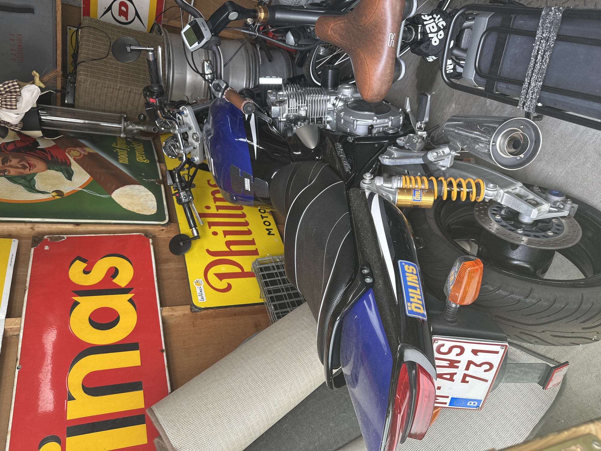 The image depicts a motorcycle parked in a cluttered garage or storage area. The motorcycle is predominantly black and blue, with a visible "Öhlins" sticker on its rear. It has a license plate that reads "AWS 731" with a Belgian flag emblem. Other objects in the scene include rolls of carpet, various signs with text and images (one appearing to be an advertisement for "in-as" and another for "Phillips"), metal containers, and a wicker basket. A brown leather bicycle seat is also visible, attached to another bicycle that is partially obscured. The overall environment suggests an indoor space with accumulated items, likely a personal storage area. The location of Bruges, Belgium, is inferred from the Belgian license plate and flag emblem on the motorcycle. There are no people visible in the image.