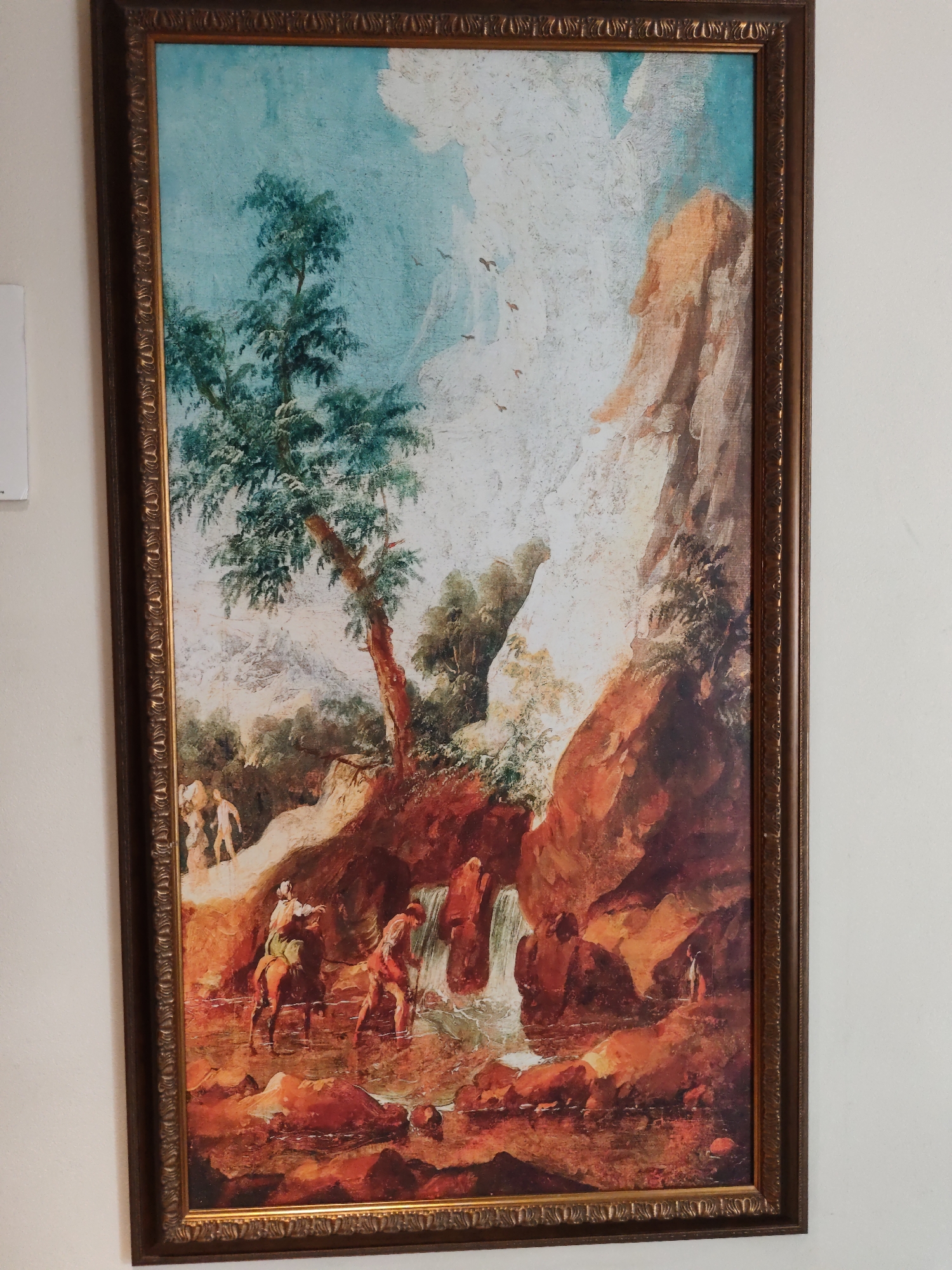 This is a framed oil painting depicting a landscape scene with figures. The painting is oriented vertically and has a dark brown, ornate frame with gold detailing.
The scene itself features a rocky, mountainous terrain rendered in warm, earthy tones of orange, red, and brown, with hints of green foliage. A waterfall cascades down the rocks into a body of water at the lower center of the painting.
Several human figures are present in the landscape. In the foreground, on the left, a figure on horseback, clad in white and green, is moving towards the right. Another figure, wearing red, is walking in the water to the right of the horse. Further back, near the waterfall, more figures are visible, some appearing to be bathing or interacting with the water. In the upper left portion of the painting, a group of figures are walking along a path. A large, mature tree with dark green foliage stands prominently in the middle left of the composition, leaning towards the right. The sky above is a bright, light blue with some white clouds and several small birds in flight.
There is no discernible connection to the city of Aihui, China, in the visual information presented in the painting or its frame.