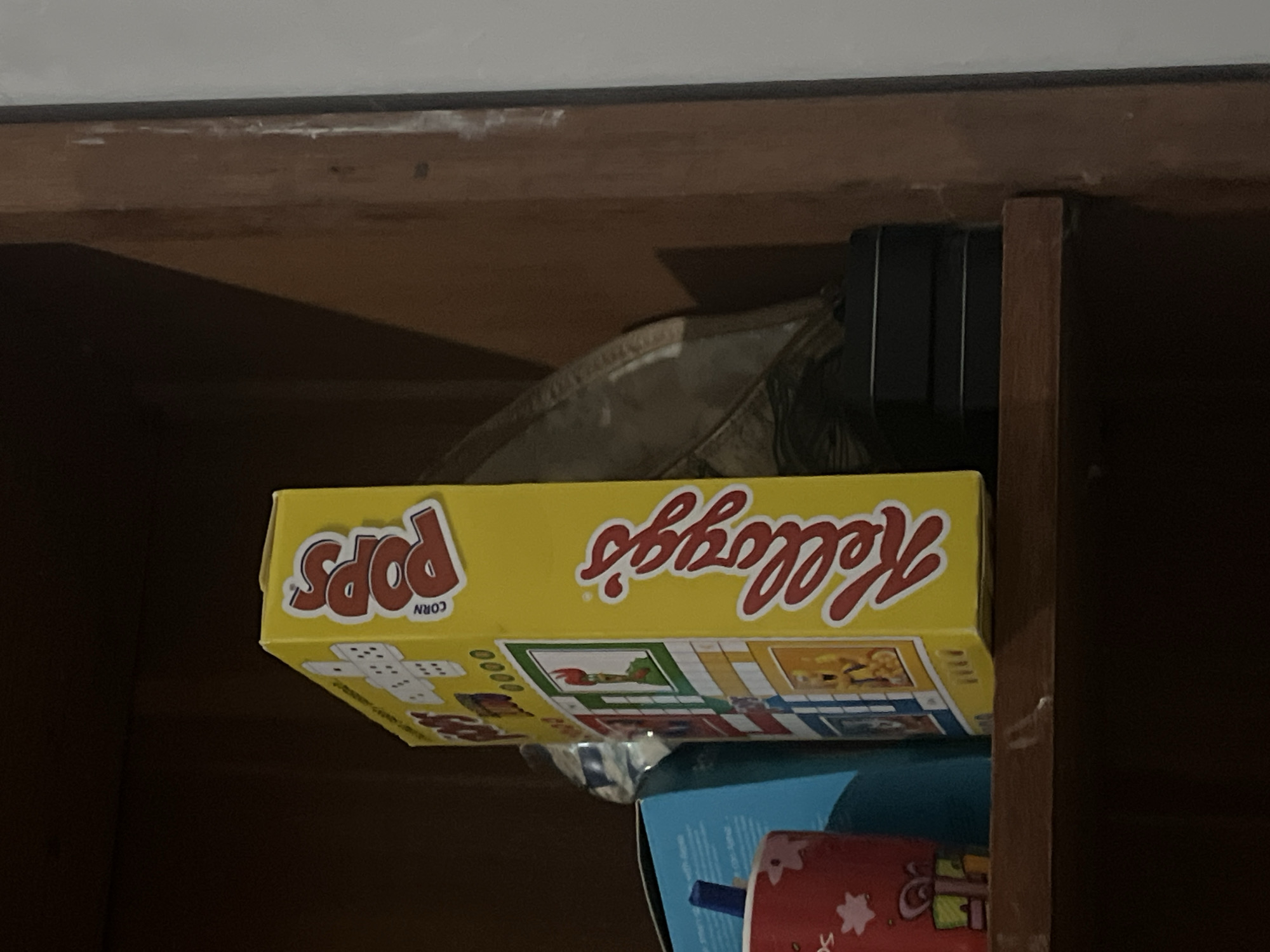 In Santa Ana, Venezuela, an indoor scene depicts a wooden shelf containing various objects. Prominently featured is a yellow Kellogg's Corn Pops cereal box, positioned vertically and facing left. To its left, a blue box and a red cup with a blue straw are visible. Behind the cereal box, a large, reflective object, possibly a platter or record, is partially obscured by shadow. At the bottom right of the shelf, a stack of three black rectangular objects rests. The shelf is set against a dark wooden background.