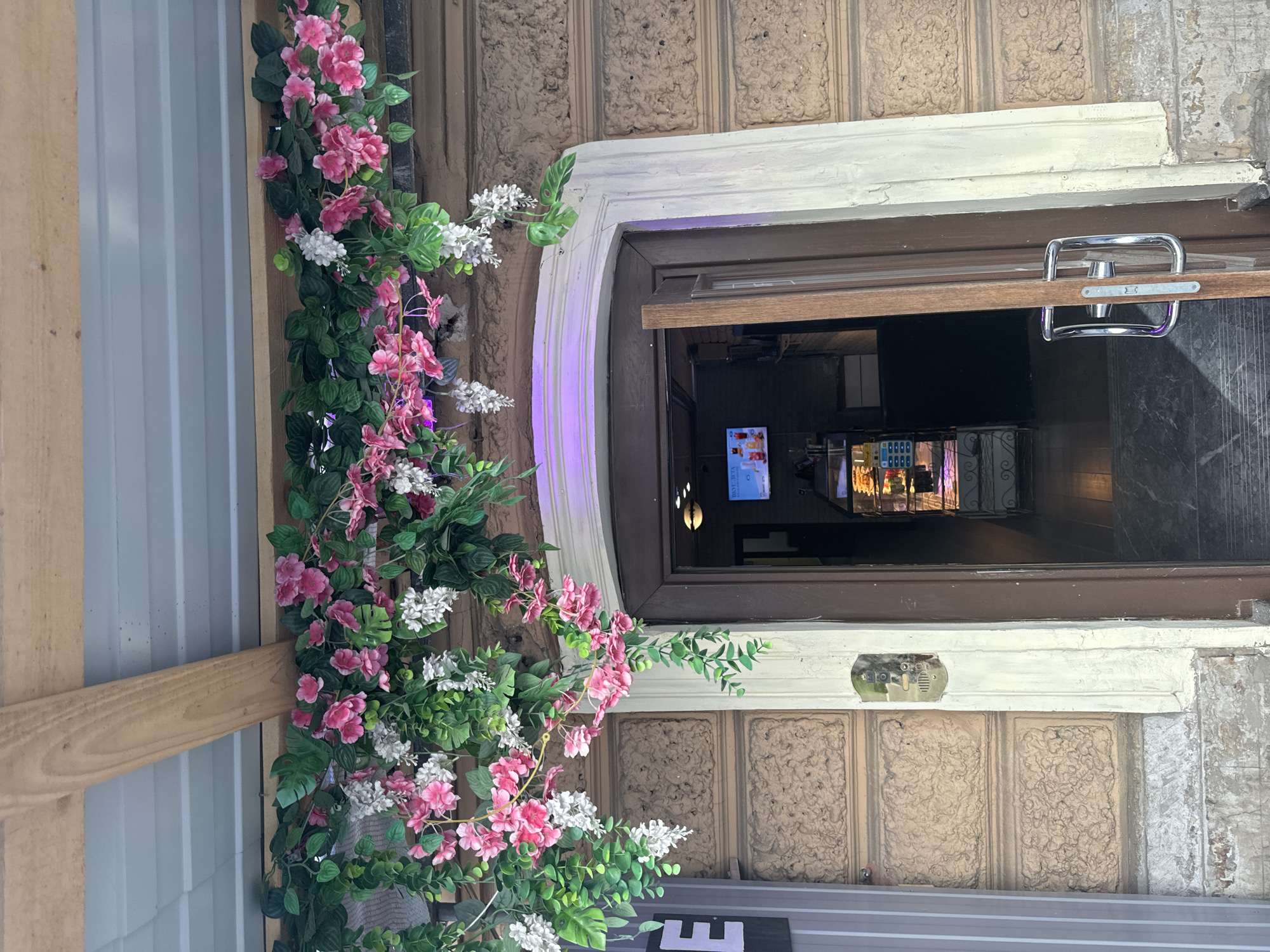 The image shows the entrance to a building in Saint Petersburg, Russia. The entrance is adorned with artificial pink and white flowers and greenery, draping over a wooden beam and the entryway. The building's facade is made of textured brown stone blocks. The doorway is a dark brown wooden door, slightly ajar, revealing a display case with baked goods inside and a television screen showing a menu or advertisement. A silver door handle is visible on the door. To the left of the entrance, a grey column with a white letter "E" is visible. The ground in front of the entrance appears to be paved with dark marble tiles, with some visible brickwork at the very bottom of the image. The overall scene suggests a storefront or cafe entrance.