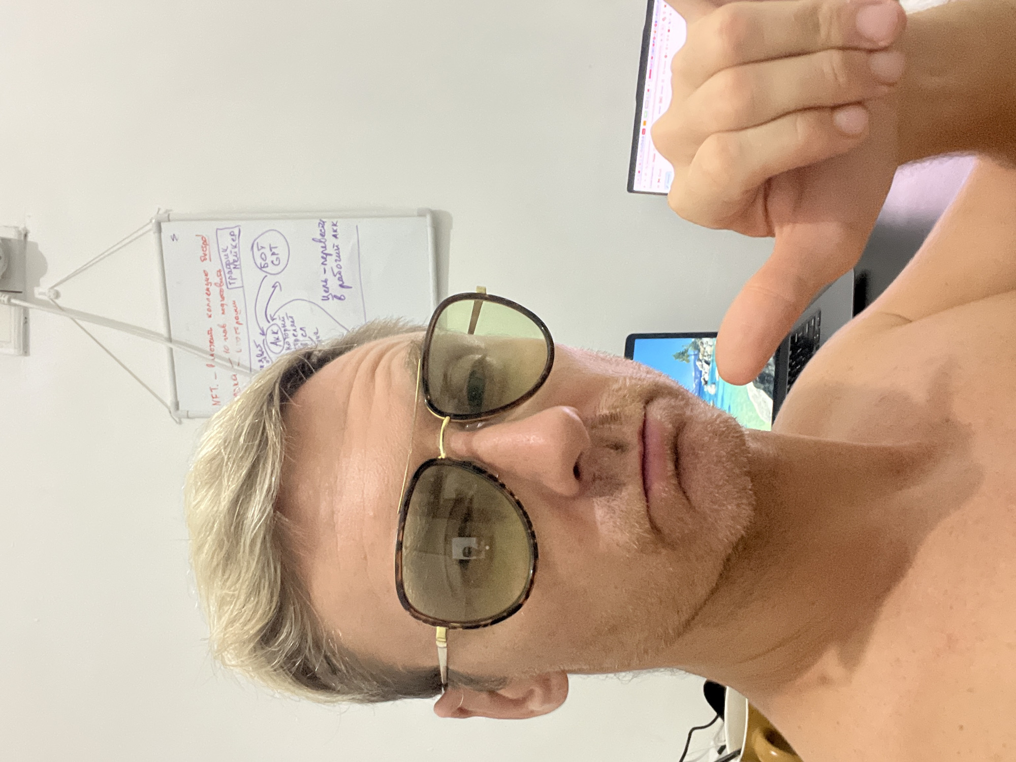 A close-up, portrait-style photo shows a man with blonde hair and aviator-style sunglasses giving a thumbs-down gesture. He appears to be shirtless. Behind him, a whiteboard with handwritten notes and diagrams is visible, along with a laptop showing a beach scene on the screen. The background includes a monitor and other items suggesting an indoor setting. The image is horizontally oriented. No location of Celuk, Indonesia can be derived from the image.