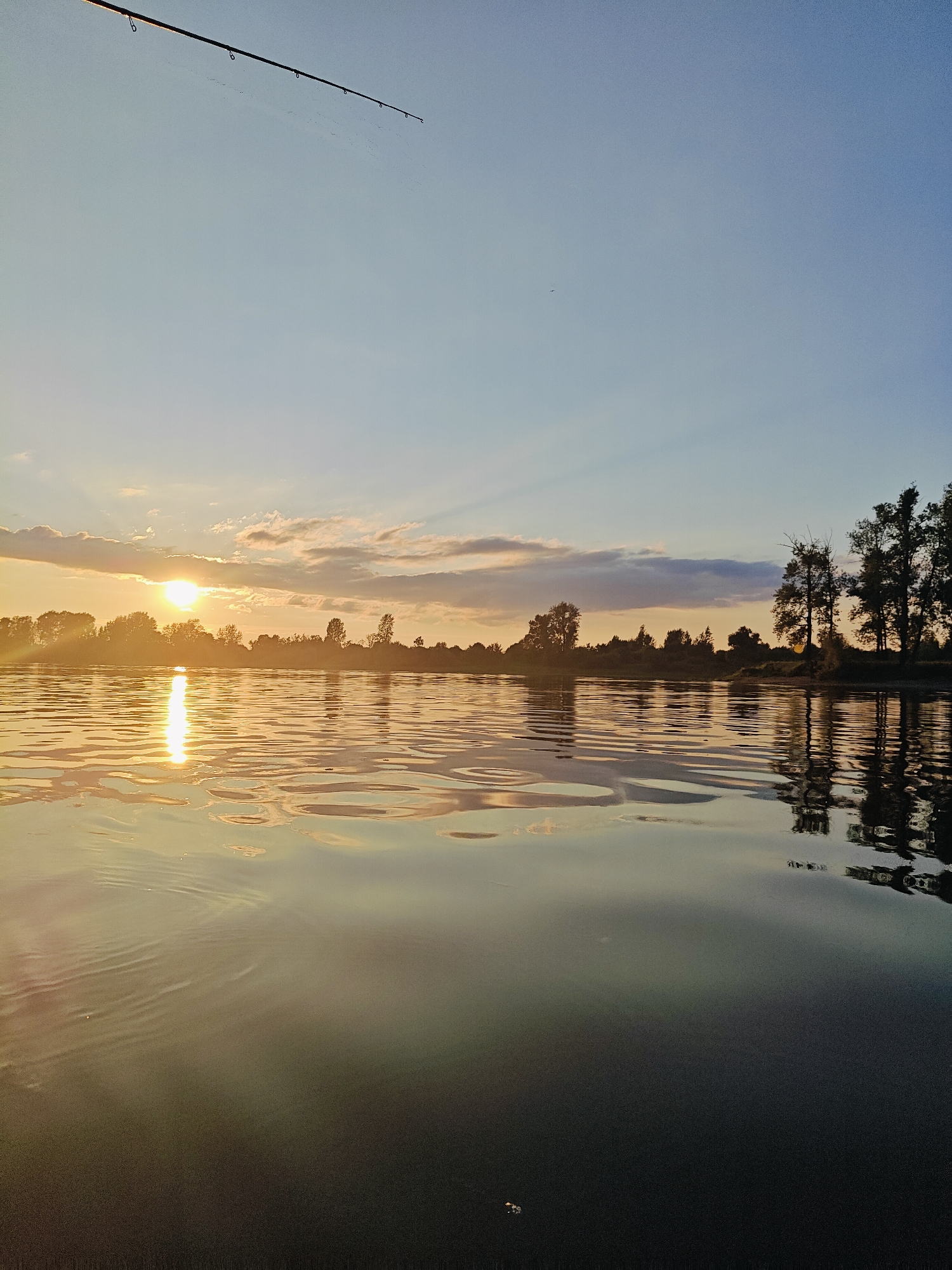 The image depicts a serene sunset over a body of water, likely a river or lake, near the village of Ol'khovskoye, Russia. The sun is low on the horizon, casting a warm, golden glow across the water and illuminating the silhouetted trees lining the distant shore. The sky is a gradient of soft blues and oranges, with scattered clouds catching the light. In the foreground, the calm water reflects the colors of the sky and the trees, with gentle ripples disturbing the surface. A fishing rod is visible in the upper left corner of the frame, angled downwards towards the water, suggesting someone is actively fishing. The overall atmosphere is one of peaceful tranquility, capturing the beauty of nature at dusk.