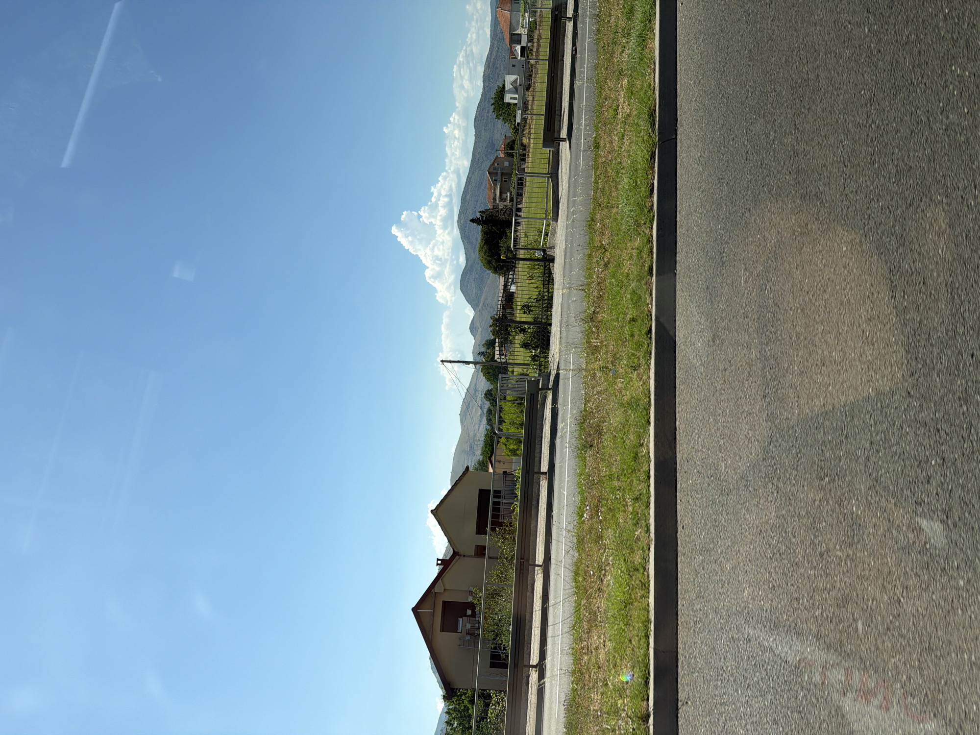 The image depicts a roadside view of a rural area, likely in or near Kruševica, Montenegro. The foreground is dominated by an asphalt road, blurred due to motion. A concrete curb separates the road from a strip of green grass, where a few small, colorful bokeh effects are visible, possibly from soap bubbles. Beyond the grass, a metal guardrail runs horizontally, delineating the edge of the road from a grassy area.
In the midground, several houses with reddish-brown tiled roofs are visible. The closest house on the left is a light-colored, two-story building with dark-colored window shutters and a balcony. Behind it, another similar house is partially obscured. Further to the right, additional houses with red roofs are scattered amongst trees and vegetation. A metal fence encloses some of the properties. In the background, a range of rolling hills and mountains under a clear blue sky with scattered white clouds forms the horizon. One distinct, fluffy white cloud is particularly prominent above the mountains. A utility pole with wires is also visible on the right side of the frame. The overall scene suggests a sunny day in a semi-rural or suburban environment.