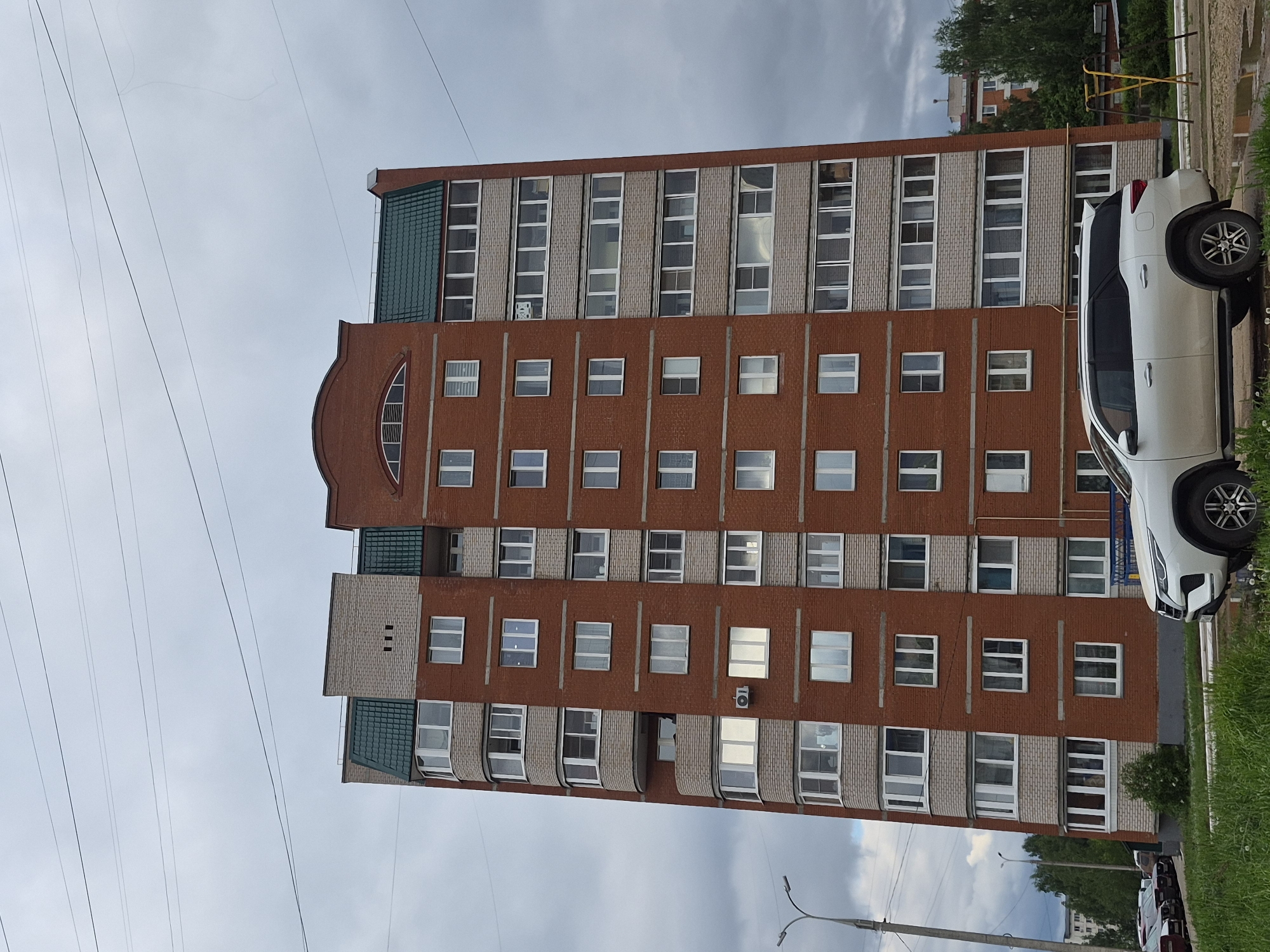 A multi-story brick building stands tall in Chaikovsky, Russia, under a cloudy sky. The building has a unique architectural design, featuring brick columns between rows of windows. A white SUV is parked on the right side of the frame, partially blocking the view of the building's facade. Green grass grows near the parked car. Power lines stretch across the sky in the background.