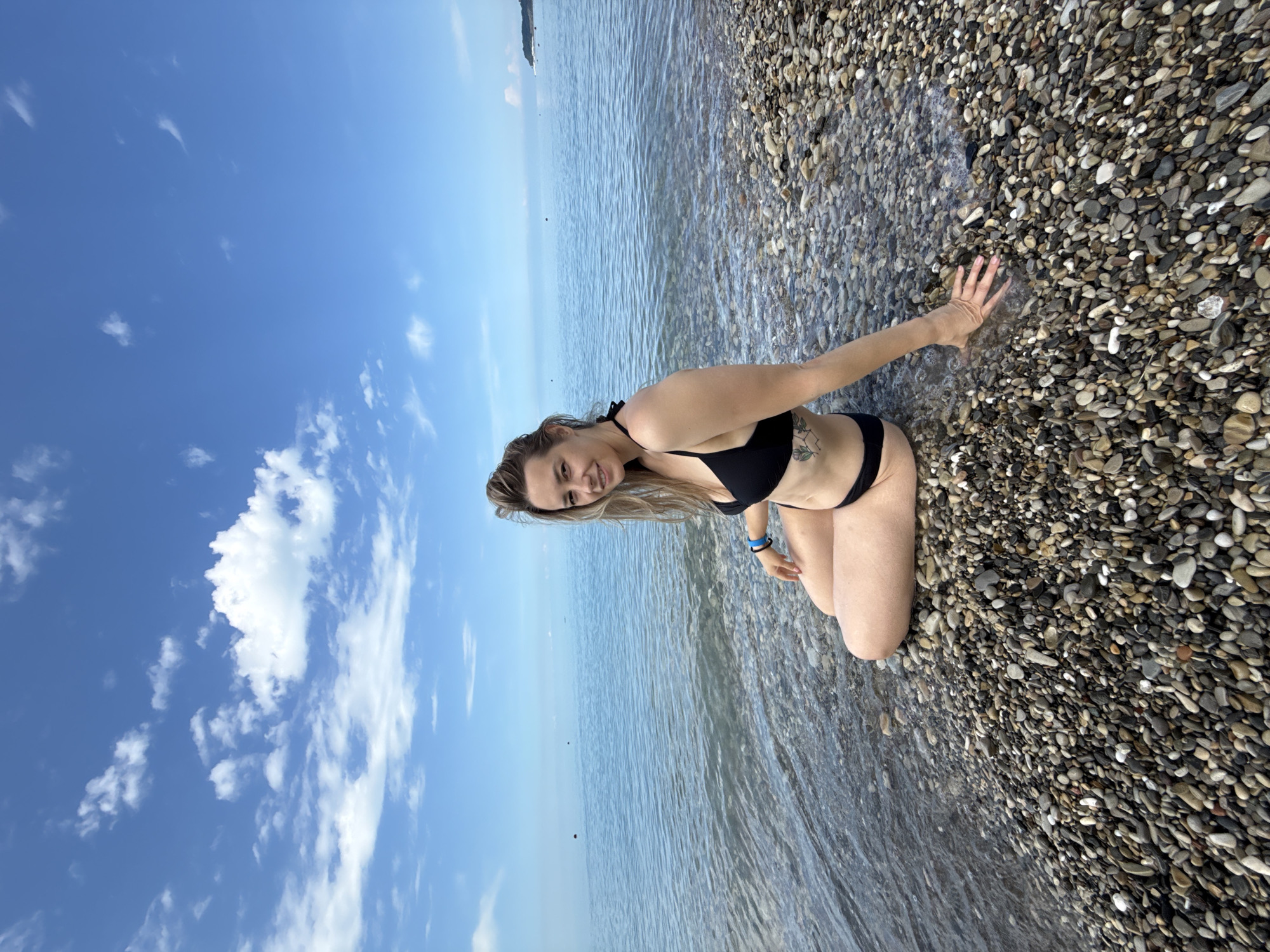 A woman in a black bikini sits on a pebble beach with the Black Sea in Nebug, Russia. The sky is blue with white clouds. The water is calm and clear. The woman is smiling and looking at the camera. Her hand is extended towards the water. She has a tattoo on her hip.
