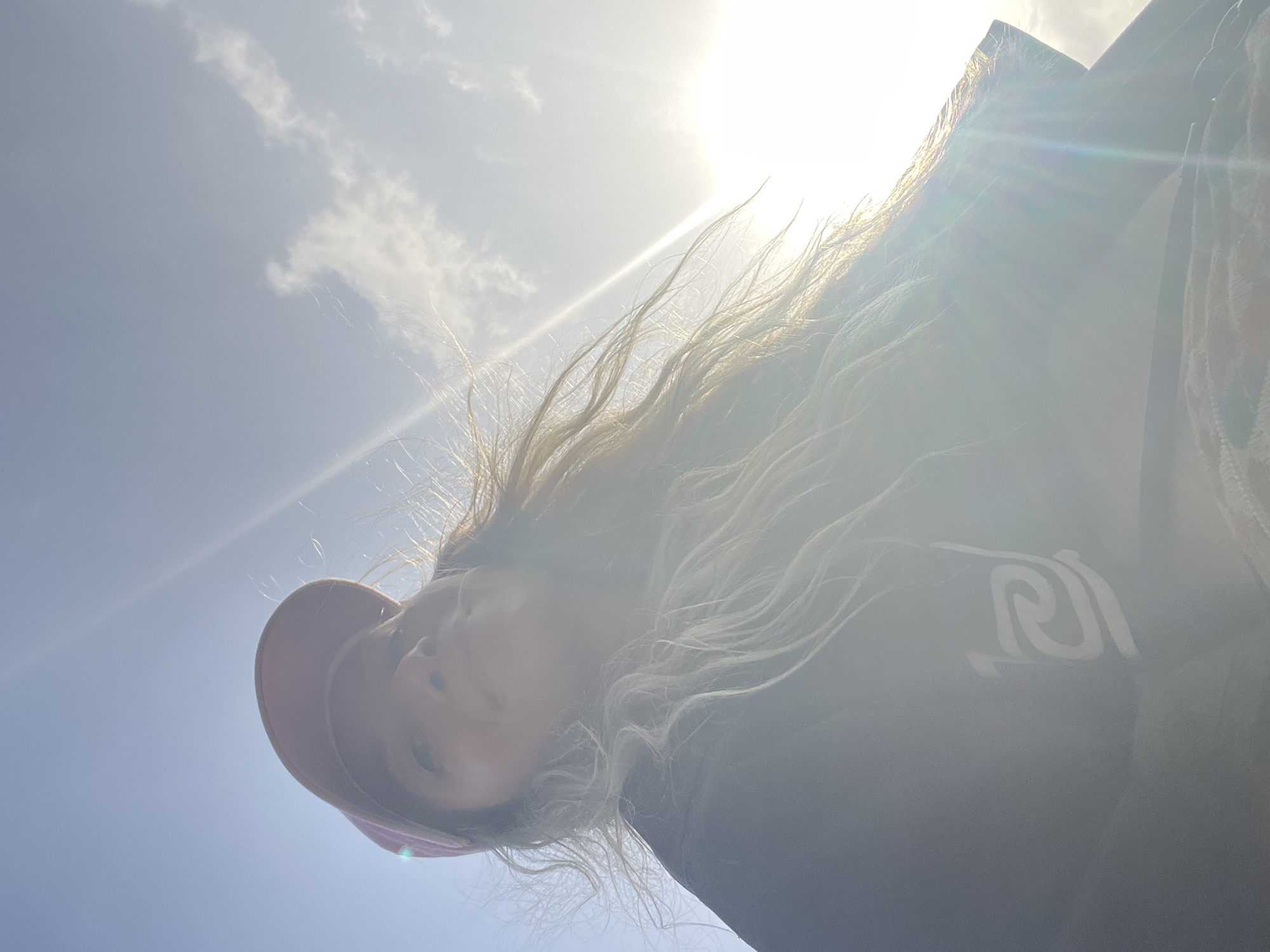 The image is a low-angle, close-up shot of a person wearing a pink baseball cap and a dark hoodie. The person's face is partially obscured by the bright sunlight, which creates lens flare across the image. Their long, light-colored hair is windswept. The background is a bright sky with a few wispy clouds and the sun positioned to the right. A white logo or graphic is visible on the hoodie. The scene appears to be outdoors, possibly in the city of Dondra, Sri Lanka, given the bright, sunny conditions typical of the region.