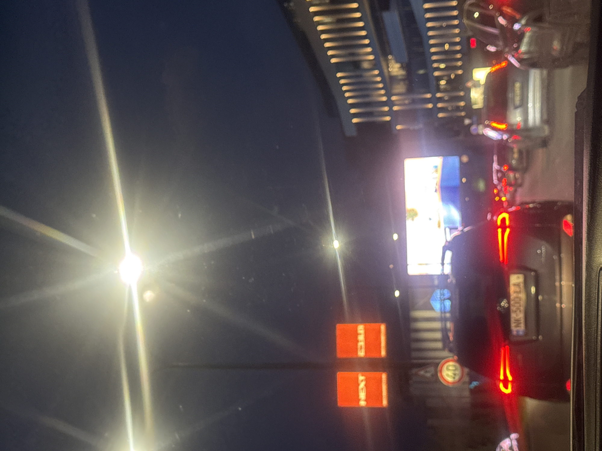The image depicts a nighttime scene in Tbilisi, Georgia, likely taken from inside a car. The view is dominated by streetlights and the illuminated signage of "NEXT" and another word, perhaps "STORE." There's a large video screen displaying content. In the foreground, the taillights of cars are visible, including one with the license plate "NK-509-ZA." A speed limit sign indicating "40" is present. The surrounding environment includes building structures with distinctive lighting. The scene suggests a busy street or thoroughfare in Tbilisi at night.