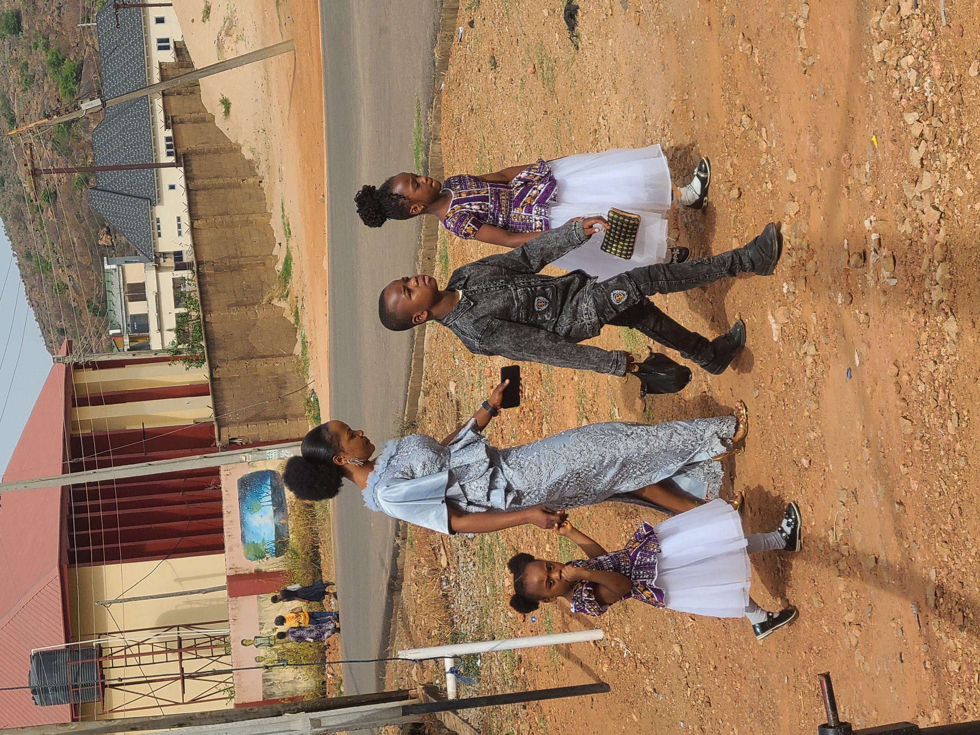 A woman, a boy, and two girls pose for a photo in Gura Top, Nigeria. The woman wears a blue dress and holds a phone, grasping the hand of a young girl in a purple and white dress. A boy in a gray denim outfit stands next to them, holding a phone. Another girl in a similar purple and white dress stands nearby, holding a clutch. The group stands on a dirt and gravel area next to a paved road. A building with a mural is visible in the background. A mountain range is visible in the distance.