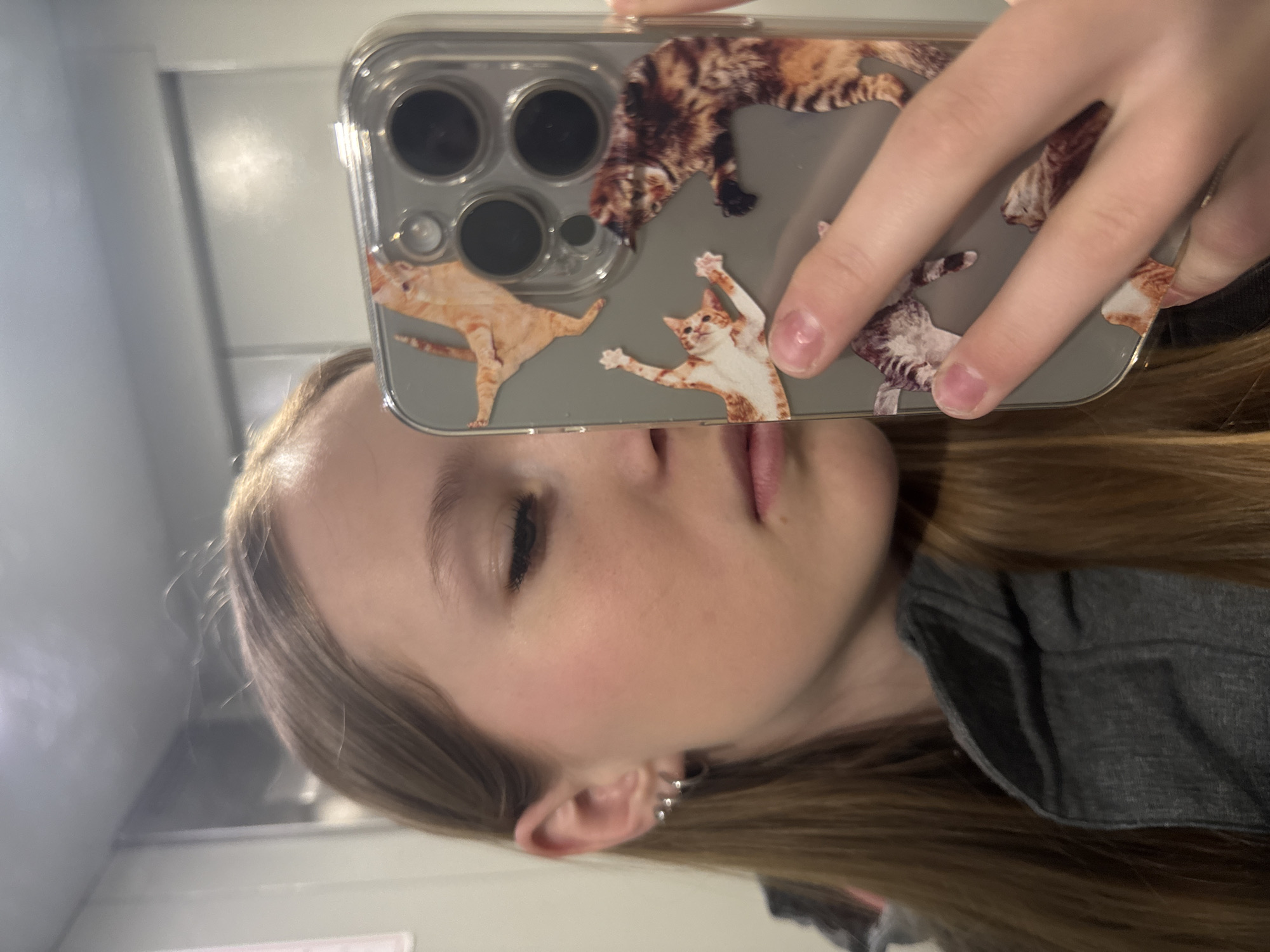 A person is taking a selfie in what appears to be an indoor setting. They are holding a smartphone with a clear case decorated with various cat images, partially obscuring their face. The person has fair skin, light brown hair, and an earring visible in their left ear. They are wearing a gray shirt. The background is nondescript, suggesting a simple interior space with plain walls. There is no indication that the photograph was taken in the city of Chaikovsky, Russia.
