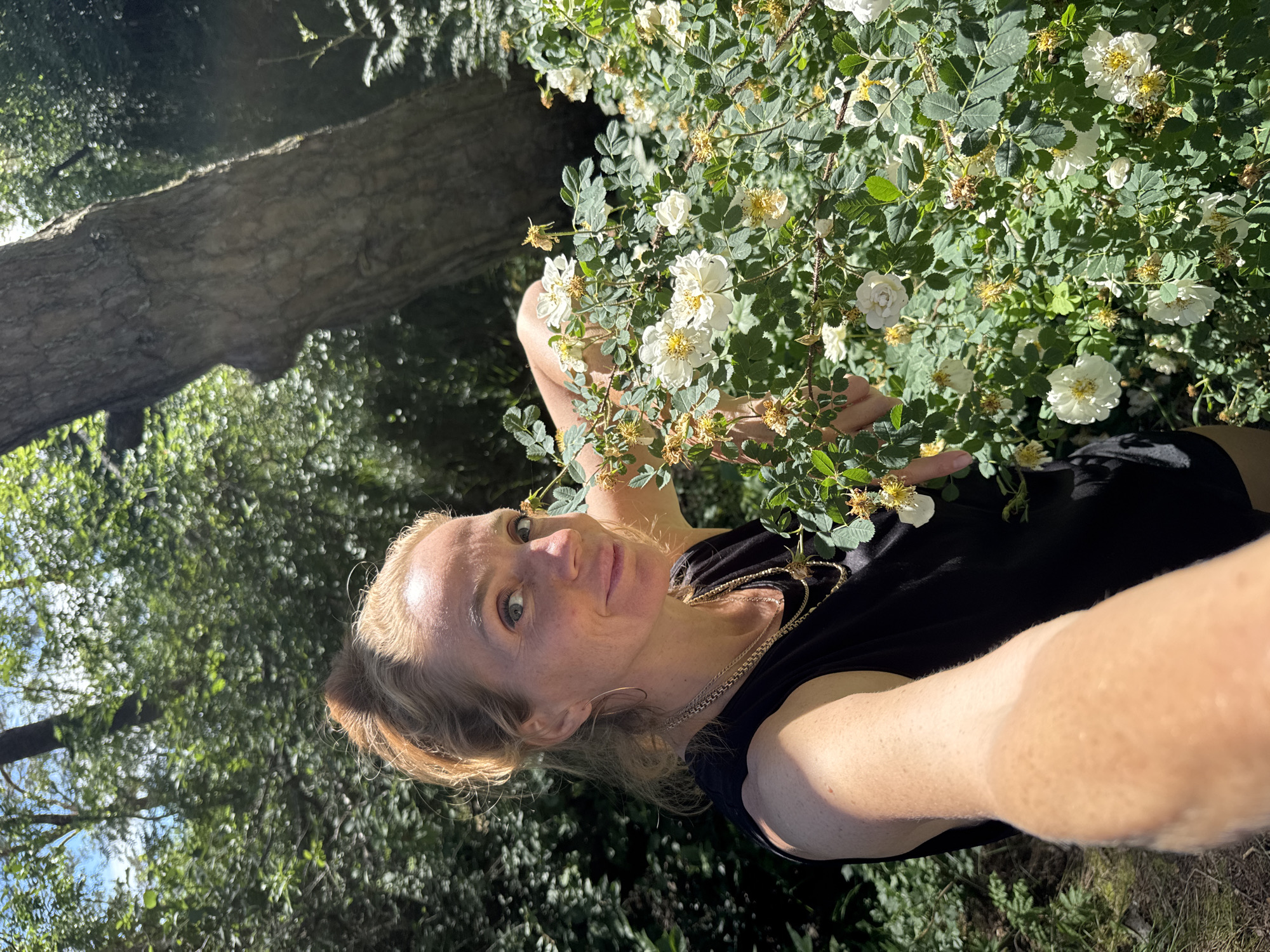 A woman with blonde hair pulled into a bun is taking a selfie in an outdoor setting. She is wearing a black sleeveless top and multiple gold necklaces. She is standing next to a bush with white flowers and green leaves. The background shows lush foliage, trees, and sunlight filtering through the leaves. The location is Zelenaya Roshcha, Russia.