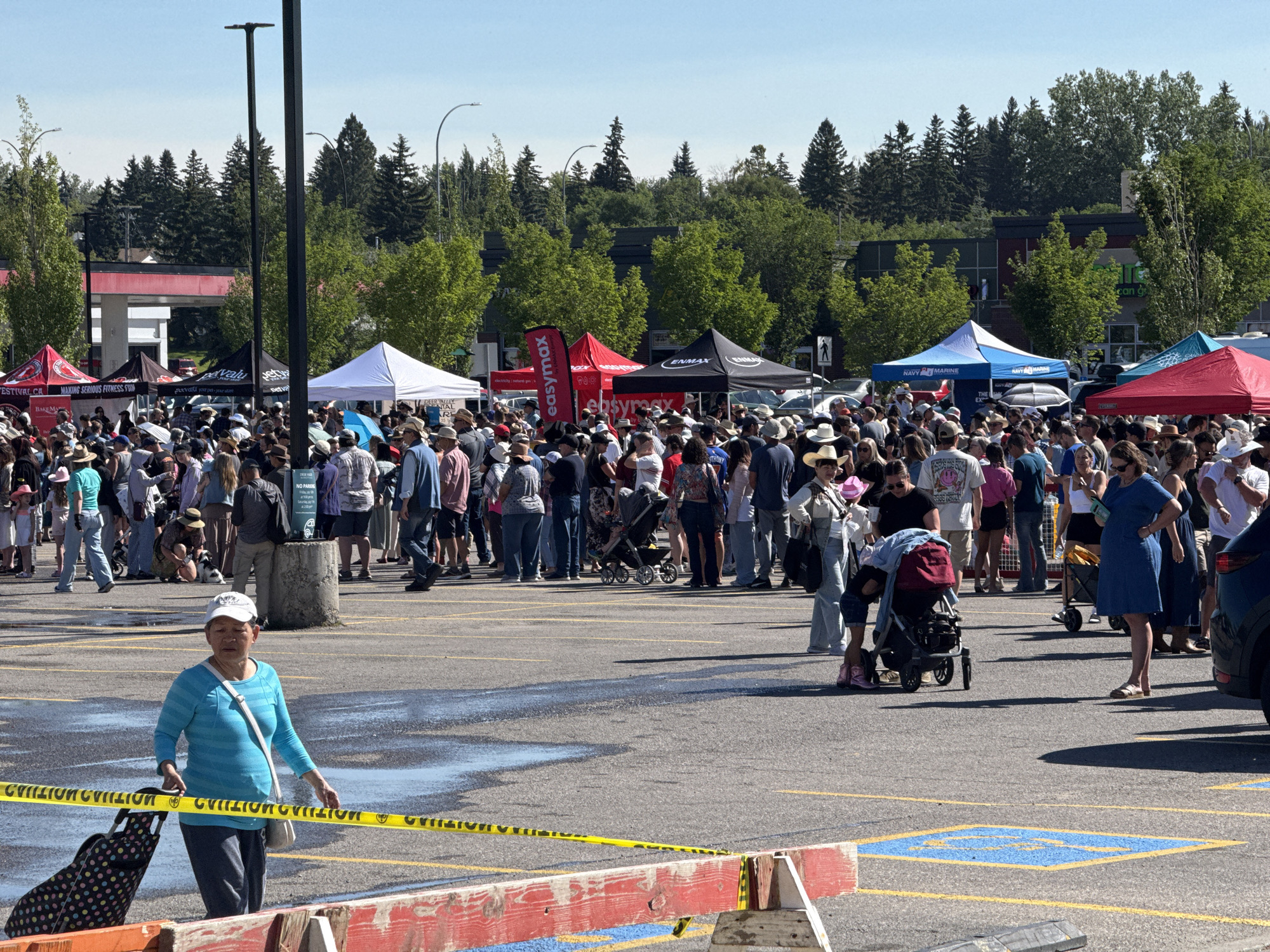 The image depicts an outdoor gathering or event in a paved parking lot under a clear blue sky. Numerous people, dressed in casual attire, are milling about, many wearing hats. Several tents, some red and white, others blue and white, are set up, suggesting vendor booths or activity areas. In the background, a row of trees borders the parking lot, and buildings can be seen behind the trees, including what appears to be a gas station with red accents and another building with dark windows.
The foreground shows yellow caution tape stretched across the pavement, along with a red and white barricade. A woman in a turquoise long-sleeved shirt and dark pants is walking with a wheeled suitcase, passing by the caution tape. Another woman in a blue dress is standing further back. The ground in the foreground has wet patches, possibly from recent rain or a spill. Parking lot lines are visible, and a blue handicapped parking symbol is painted on the asphalt in the mid-right of the frame.
Given the context of the event and typical Calgary events, this scene is likely from the parking lot of Spruce Meadows, a large equestrian facility located in Calgary, Alberta, Canada, which often hosts major events and festivals.