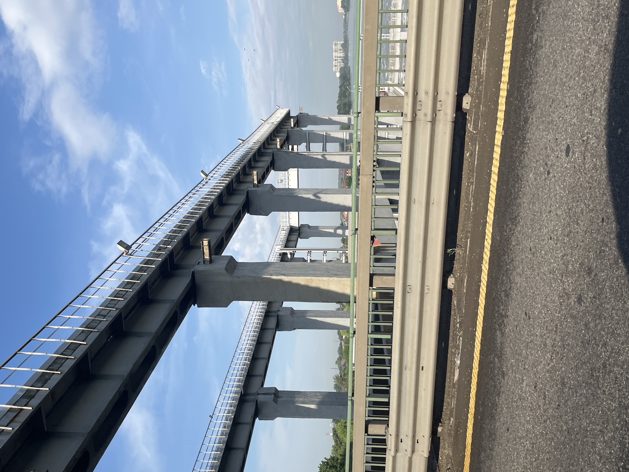 The image captures an elevated concrete bridge structure under a partly cloudy blue sky. The bridge, supported by numerous large concrete pillars, extends horizontally across the frame. A secondary, lower bridge runs parallel to the first, also supported by pillars. Both bridges feature railings along their edges. In the foreground, a road with a yellow dividing line is visible, and a metal guardrail separates the road from the bridge structure. Beyond the bridge, a body of water can be seen, with buildings and trees along its banks. The scene appears to be an urban or suburban landscape, possibly in Tlustenkhabl, Russia, given the context of the prompt. There are no visible people or animals in the image, and the overall atmosphere is one of infrastructure and a somewhat overcast day.