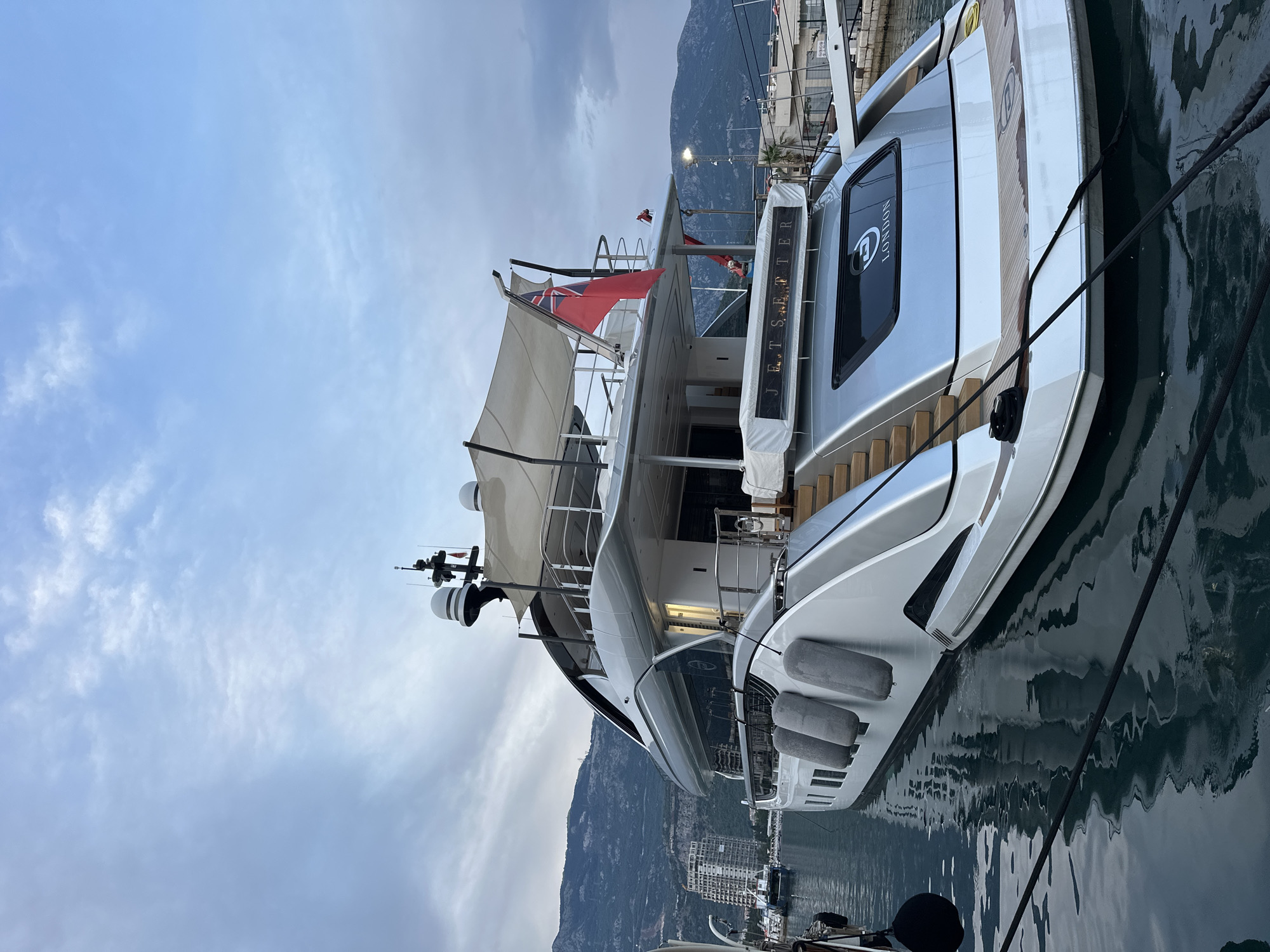 The media file shows a large, luxurious yacht docked in a harbor with mountains in the background, likely Budva, Montenegro. The yacht is predominantly silver and white, with wooden accents and dark tinted windows. The name "JET SETTER" is visible on a plaque towards the stern, along with the word "LONDON" on one of the windows. A British flag is prominently displayed on a mast.
The yacht appears to be at rest, docked with thick black ropes. There are several grey fenders hanging along its side to protect it from the dock. The water is dark and reflects the yacht and the overcast sky, indicating either early morning or late evening, or a generally cloudy day. The lighting suggests it's not a bright, sunny day. The overall atmosphere is one of quiet elegance and opulence. No people are clearly visible in the media file. The scene is a harbor, likely a marina, given the presence of multiple boats and docking facilities. The mountainous backdrop is a prominent feature of the landscape, characteristic of the Adriatic coast.