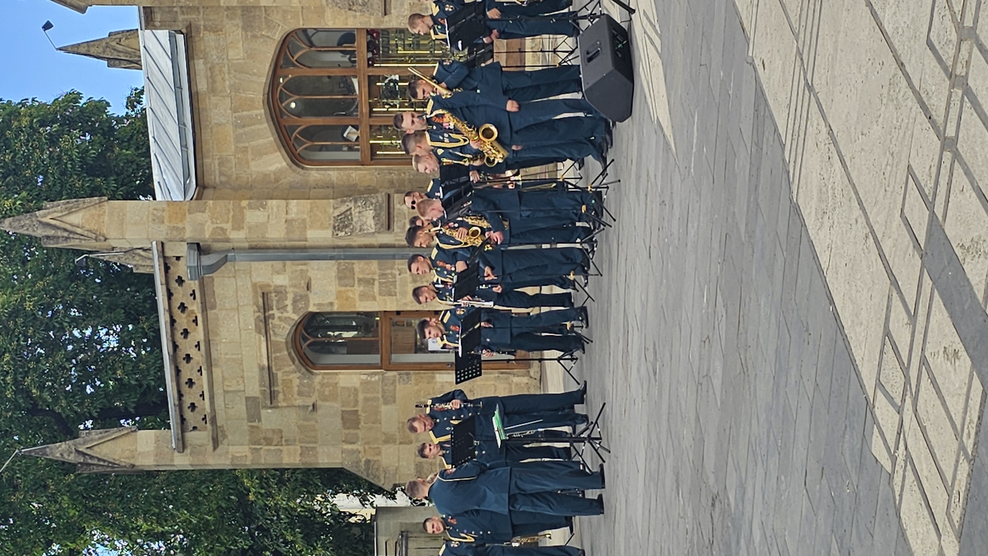 A military band is performing in front of a stone building, which is the Narzan Gallery in Kislovodsk, Russia. The band members are wearing dark blue uniforms with gold trim and medals. They are playing various instruments, including saxophones, clarinets, and what appears to be a flute or piccolo. The building has gothic-style architecture with arched windows and ornate stonework. The ground in front of the building is paved with large stone tiles. There are music stands set up for the musicians, and a black speaker is visible in the foreground. The sky is clear and blue.