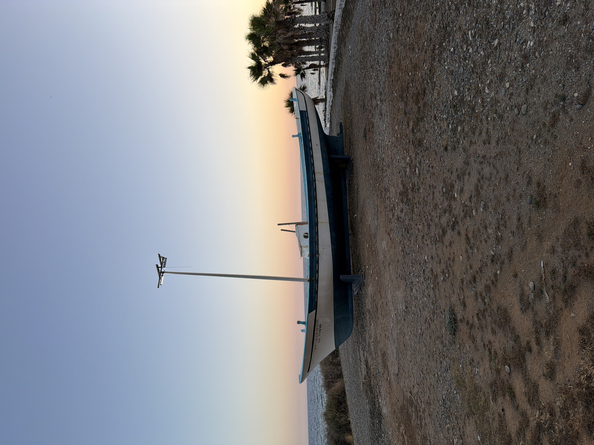The image displays a stationary traditional fishing boat, locally known as a *kaiki*, positioned on an unpaved, gravelly ground at the coastal area of Pervolia, Cyprus. The boat is painted white with a prominent teal blue lower hull and trim, appearing weathered but intact. The bow features the Greek inscription "ΑΓ. ΕΙΡΗΝΗ" (Ag. Eirini, meaning Saint Irene) and "LL 956." A tall, metal street light pole extends vertically from the center of the boat, equipped with two horizontal light fixtures, one of which is illuminated. The boat rests on dark-colored support stands.
To the left of the boat, the Mediterranean Sea is visible with gentle waves breaking on a low, rocky, and sparsely vegetated coastline. To the right, a cluster of palm trees stands near the water's edge, partially obscuring the sea, with a low concrete barrier or path running horizontally. The ground in the foreground and midground consists of dry, reddish-brown earth mixed with various sizes of gravel and sparse patches of dry vegetation.
The sky above exhibits a clear, gradient transition, ranging from a pale blue at the zenith to soft hues of peach and light orange near the horizon, indicative of either sunrise or sunset. No individuals are present in the scene.