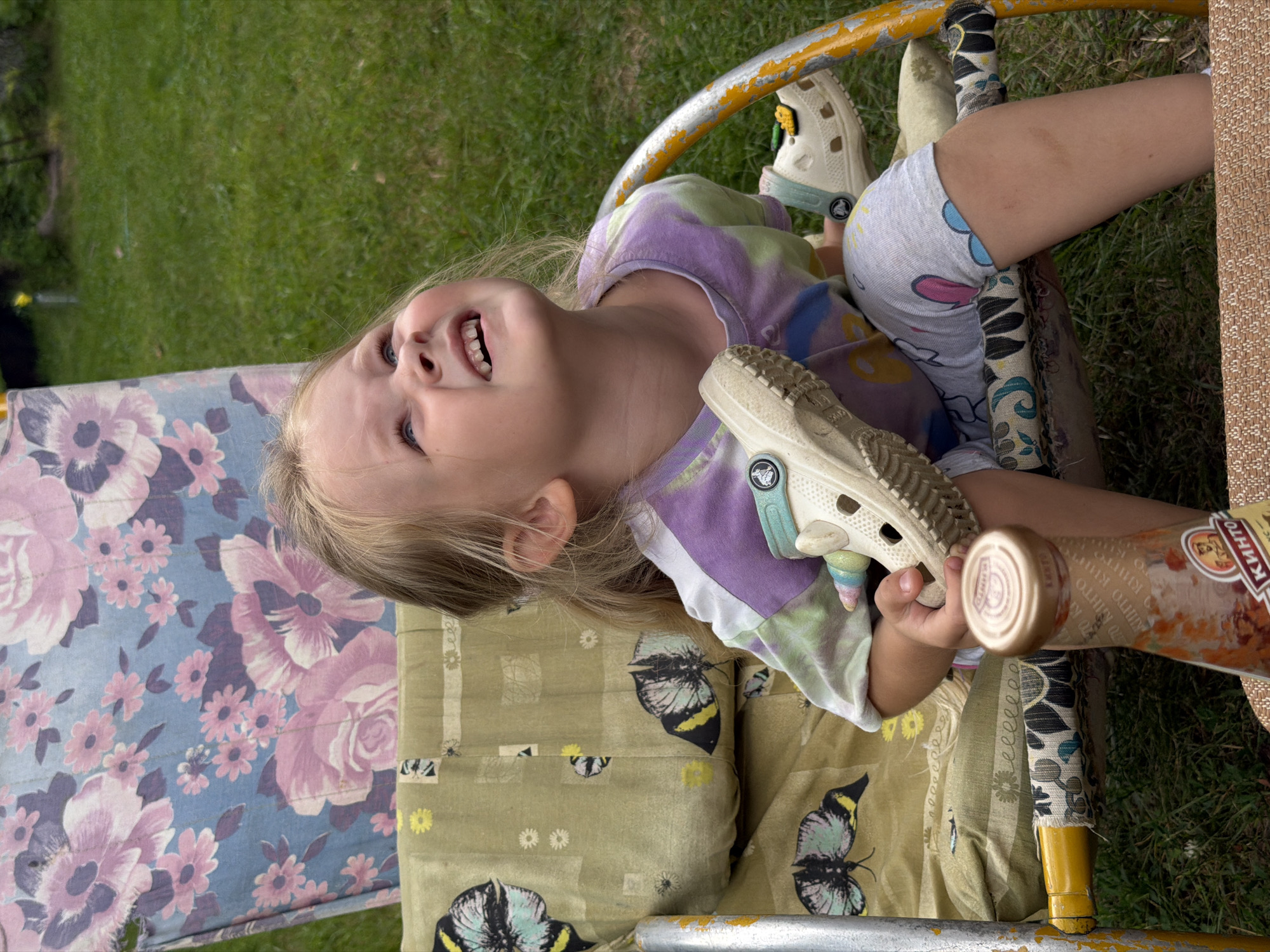 A young Caucasian girl with blonde hair, wearing a tie-dye t-shirt and patterned leggings, sits in a weathered outdoor chair in a backyard in Udelnaya, Russia. She is looking upwards and to her right, with an amused expression. Her right hand is holding a beige Croc shoe, and a partial bottle of what appears to be ketchup or a similar condiment stands on a table in the foreground. The chair has a floral patterned fabric on the backrest and a green fabric with butterfly designs on the seat. The background consists of green grass and foliage, typical of a residential outdoor setting.