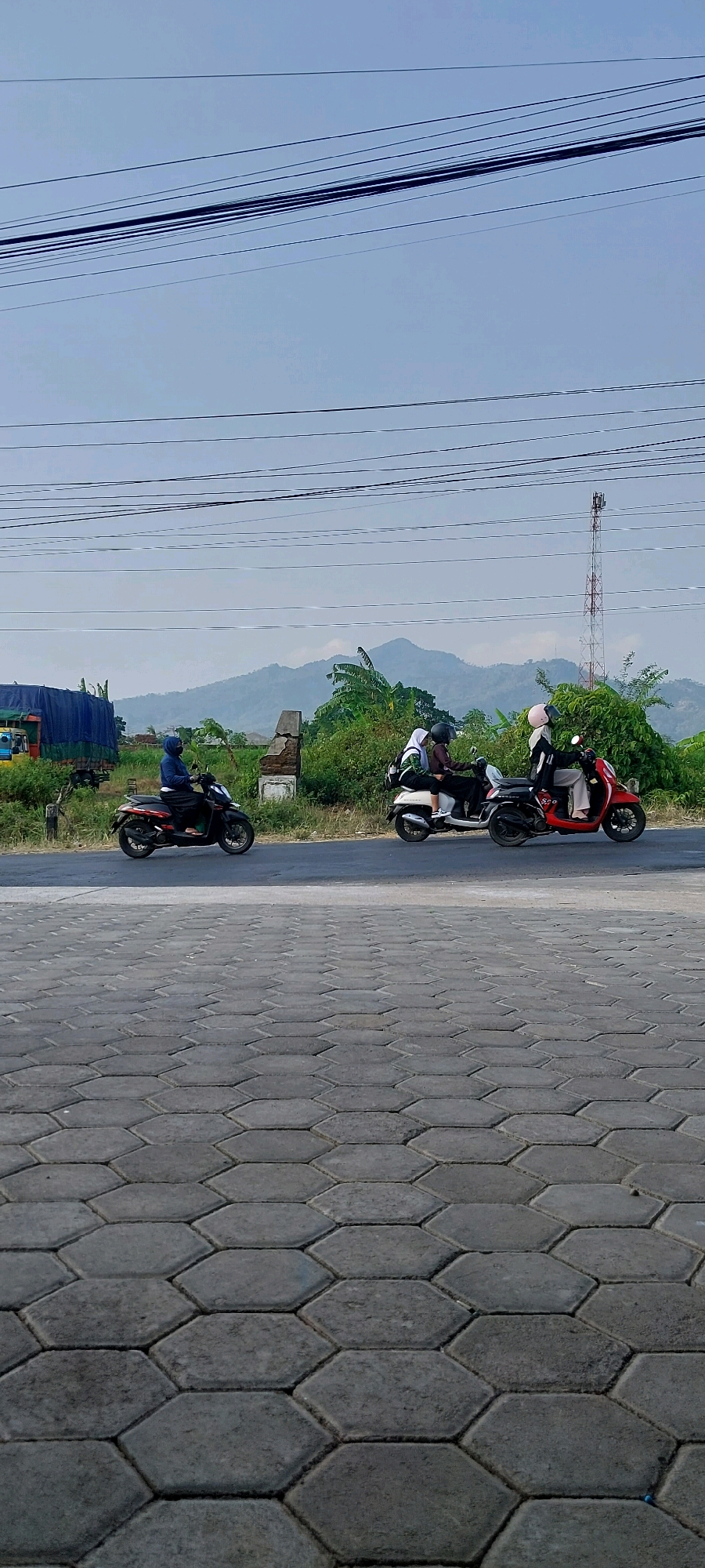 The image shows a street scene in Keling, Indonesia. In the foreground, a hexagonal patterned pavement leads to a paved road where three motorcycles are traveling. The first motorcycle, a black scooter, is ridden by a person wearing a blue hooded jacket and a black mask. Behind them, a white scooter carries two people. The rider of the white scooter wears a white hijab and a green and black jacket. The passenger on the white scooter wears a white hijab and a dark jacket. The third motorcycle, a red scooter, is ridden by a person wearing a white hijab and a black jacket. In the background, there are trees, a large blue tarp-covered truck, a damaged stone structure, and a telecommunications tower. The scene is set against a backdrop of distant mountains and a hazy sky. The overall impression is one of daily life in a semi-urban or rural Indonesian setting.