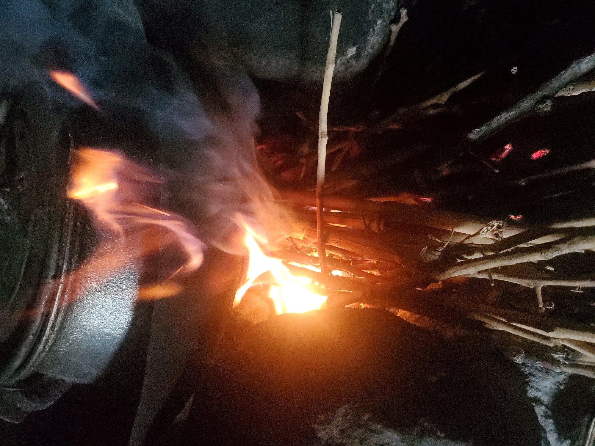 The image shows a close-up of a cooking fire. A large black pot is positioned above a fire pit, with flames licking upwards. The fire is made of a pile of sticks and branches, some of which are glowing red. Smoke is rising from the fire and obscuring parts of the scene. The background appears to be a dark, possibly rocky, environment. The overall impression is one of a rustic and traditional cooking setup. The location is unknown.