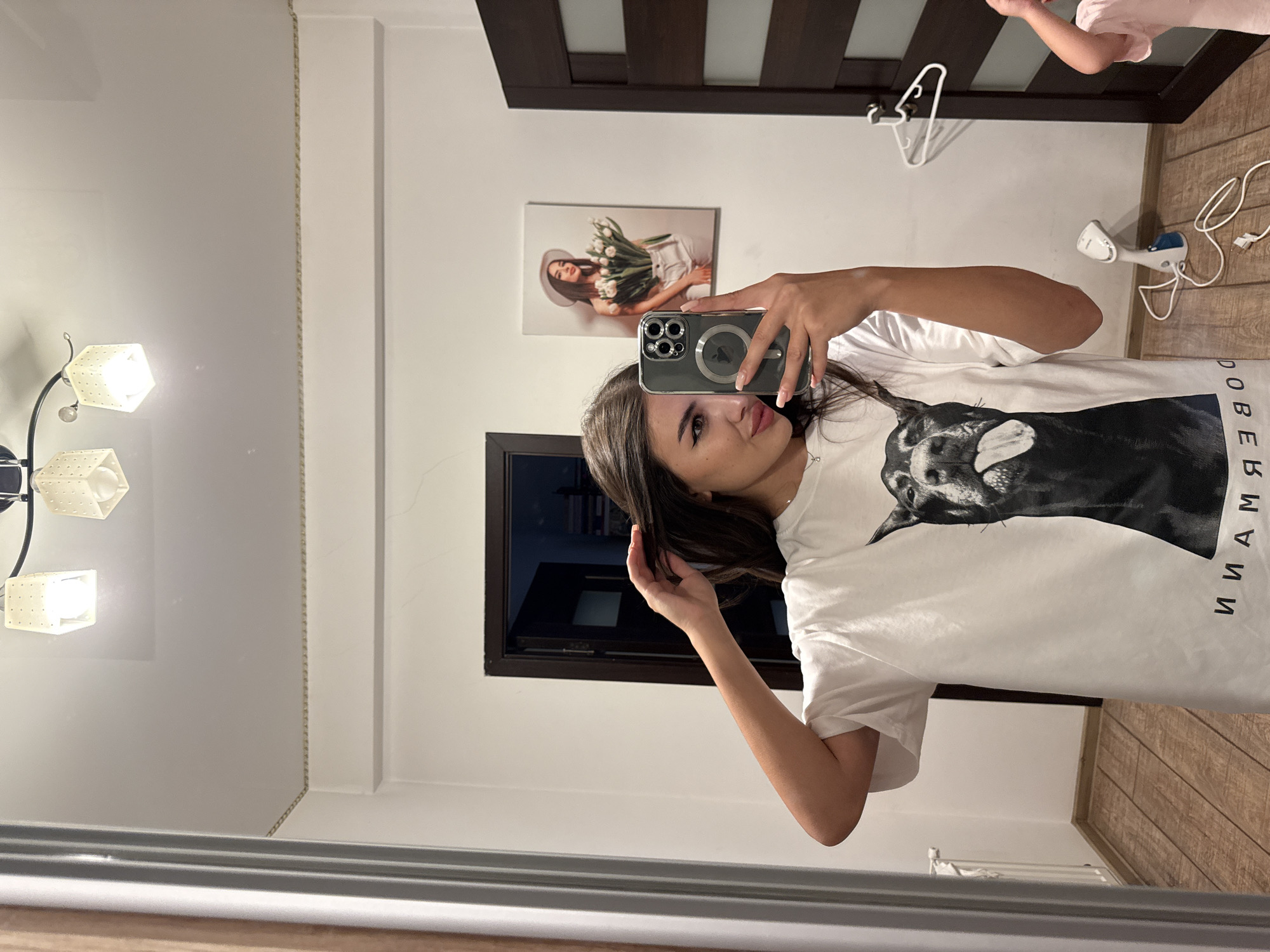 A young woman is taking a mirror selfie in what appears to be a hallway or dressing room. She is wearing a white t-shirt with a large graphic of a doberman dog sticking its tongue out and text below it. She has long, dark hair, is wearing subtle makeup, and is holding her phone up to take the picture. In the background, there is a door, a framed picture of a woman holding tulips, a light fixture on the ceiling, and a handheld garment steamer on the floor. The floor is made of light-colored wood planks. There is no indication of the specific location within Chișinău, Moldova.