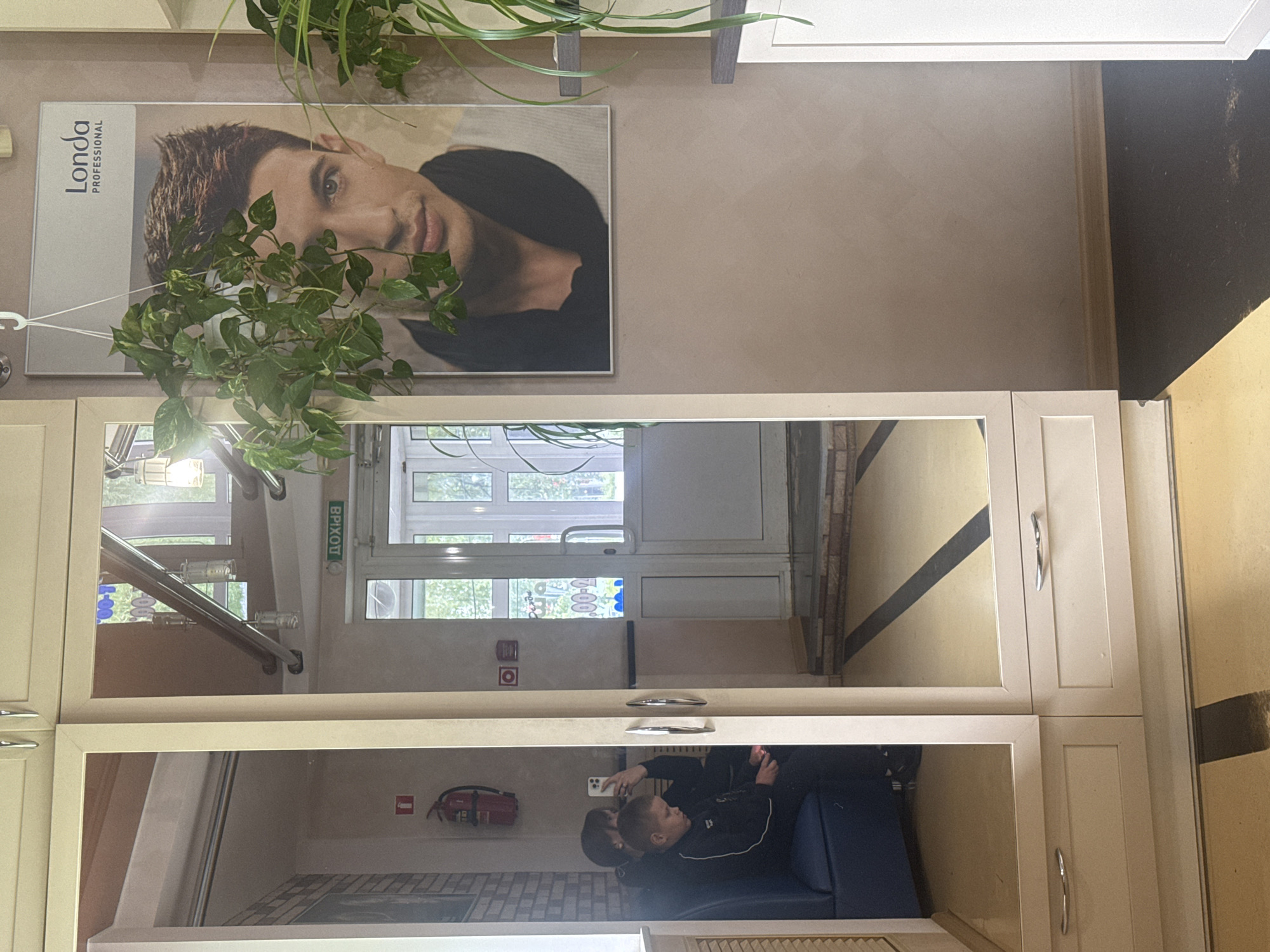 The image shows a reflection in a mirrored cabinet. In the reflection, a room is visible with a door leading to the outside. An emergency exit sign written in Russian is near the door. There is a staircase visible and a fire extinguisher mounted on the wall. Two people, seemingly a child and an adult, are sitting on a blue couch, with one of them holding a smartphone. The mirror is surrounded by a light-colored cabinet. Above the cabinet is a promotional poster for Londa Professional. A potted plant hangs above the poster. The location of the image appears to be indoors, possibly a waiting area or a salon in the city of Chaikovsky, Russia.