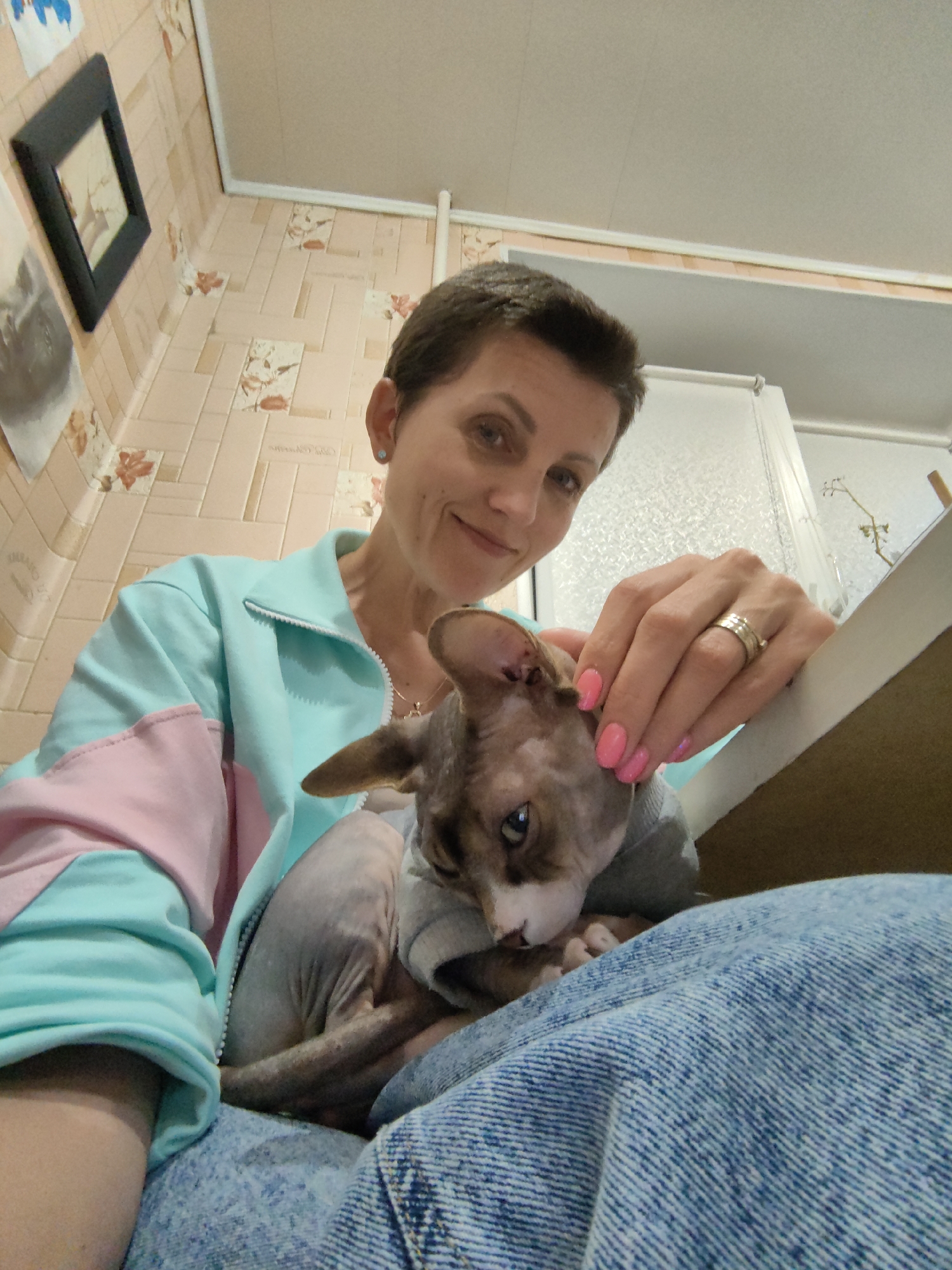 A woman with short brown hair is holding a Sphynx cat while sitting on a surface covered with blue denim material. She is wearing a light blue zip-up jacket with pink accents on the sleeves and a necklace. Her right hand, adorned with rings and bright pink nail polish, rests on the cat's head. The cat is wearing a grey garment. The background features light-colored tiled walls with a floral pattern and a white ceiling. A framed picture is visible on the wall to the left. The scene appears to be indoors, possibly in a residential setting. The location is the city of Myadzyel, Belarus.