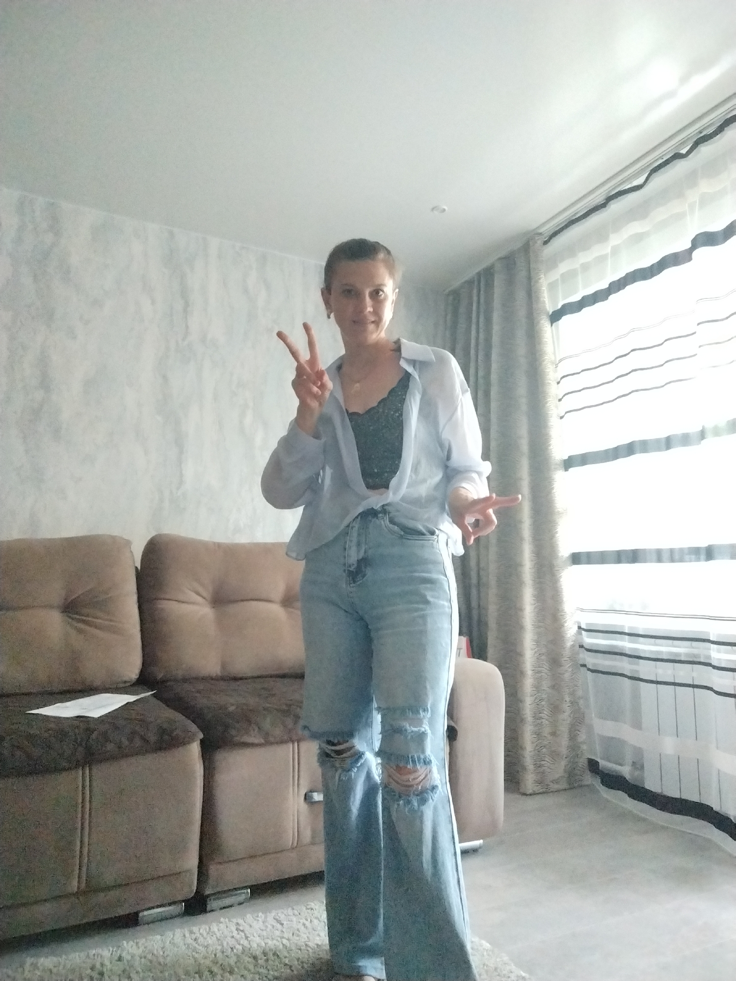 A young woman stands in a living room, posing for a photo. She is wearing a light blue unbuttoned shirt over a dark lace bralette, and light wash ripped jeans. She has her hair pulled back in a bun and is making a peace sign with her right hand, while her left hand is extended outwards, palm facing forward. The room features a brown sectional sofa with dark brown accents, a textured grey wall, and a window with striped curtains. A small white envelope rests on the sofa cushion. The floor is covered with a light-colored rug and grey flooring.