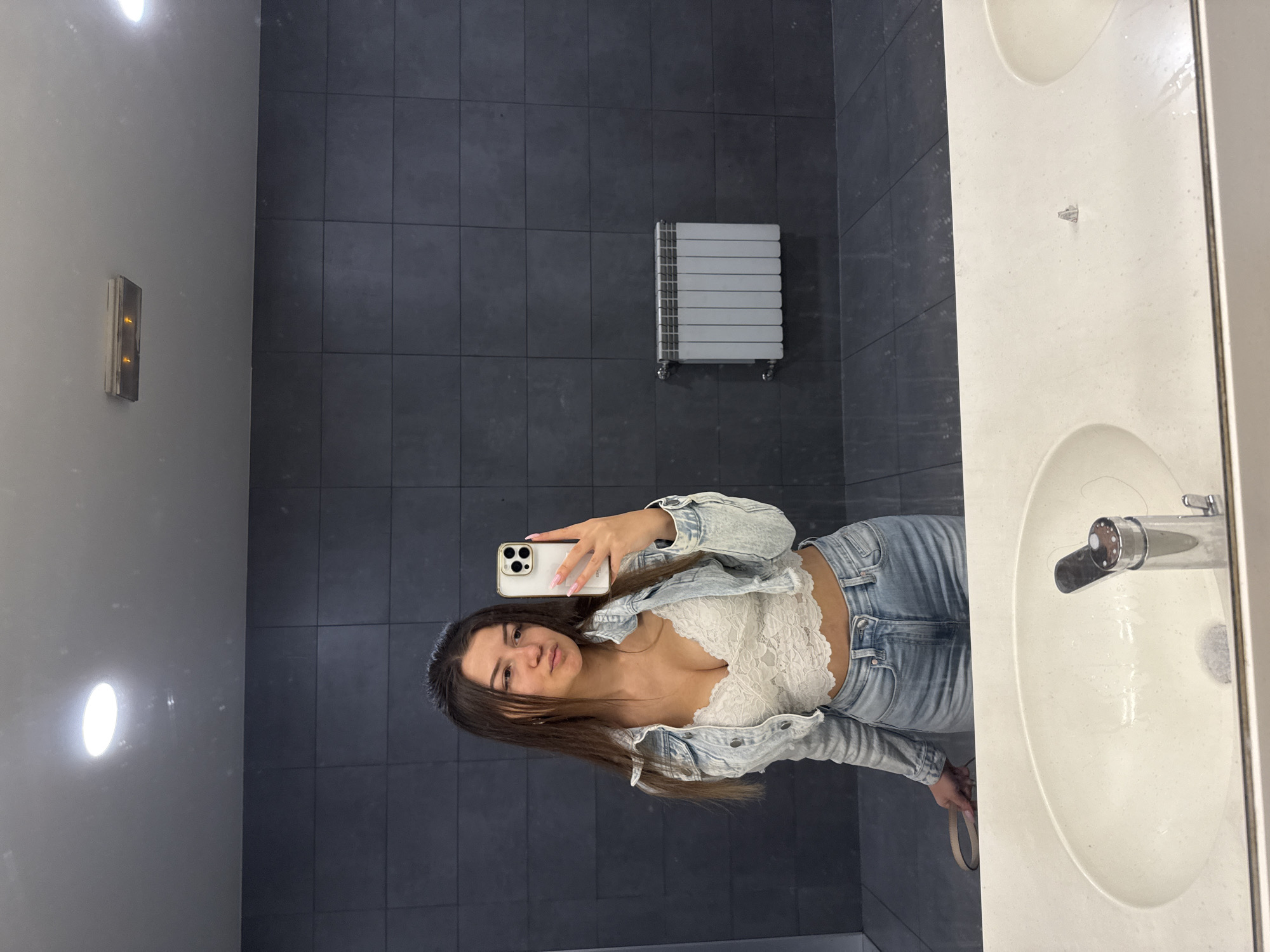 A woman is taking a selfie in a bathroom. She is wearing a white lace top, a denim jacket, and jeans. The bathroom has gray tiled walls, a white radiator, and a white sink. There is a light fixture on the left wall. The location is unknown and could not be confirmed as Sofia, Bulgaria.