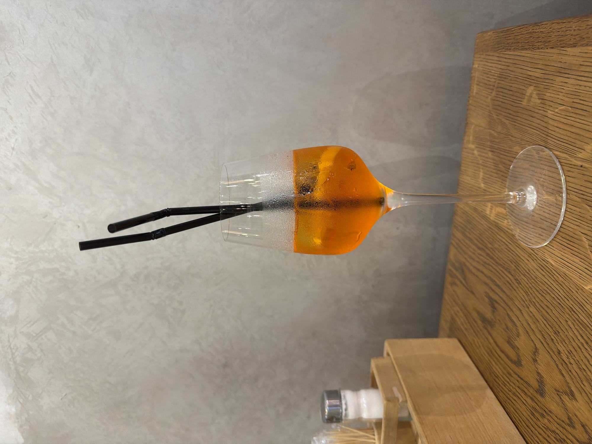 A high-angle, medium shot captures a bright orange cocktail with ice and an orange slice, served in a tall stemmed wine glass. Two black straws, one slightly longer than the other, are inserted into the drink. The glass is sweating, indicating it is cold. The cocktail is positioned on a wooden table with a visible wood grain pattern. To the left of the cocktail, a wooden holder contains a salt shaker and toothpicks. In the background, a concrete wall with a textured finish is visible. The scene suggests a casual dining or bar setting. The location is identified as Бараўлянскі сельскі Савет, Belarus.