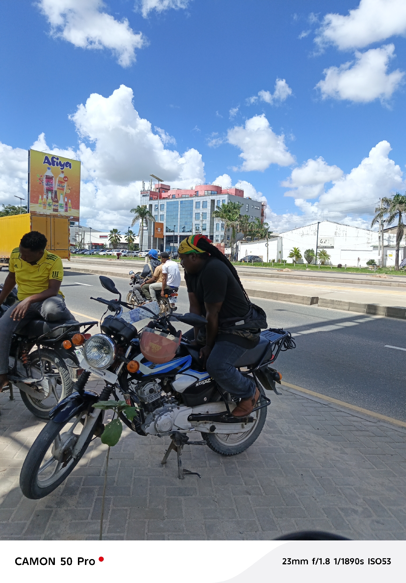 Here's a concise, forensic description of the image:
**Scene:** A street scene in Dar es Salaam, Tanzania, on a sunny day with scattered clouds. The image is taken from a ground-level perspective.
**People:** Three men are visible. Two are sitting on motorcycles parked on the sidewalk alongside the road. One man wears a yellow shirt and grey pants. The other man wears a black shirt and blue jeans, with his hair in dreadlocks and a Rasta headband. A third man, clad in a white shirt, rides a motorcycle further down the road.
**Objects:** The dominant objects are the two motorcycles parked on the sidewalk. A billboard displaying "Afiya" beverages is visible in the background, along with buildings, one possibly labeled "Makonda House." A small sapling sprouts from the sidewalk in front of one of the motorcycles. A helmet rests on the handlebar.
**Actions:** The men on the motorcycles are not actively riding, but are seemingly conversing.
**Environment:** The scene takes place on a sunny day with a partially cloudy sky. The backdrop includes urban buildings, palm trees, and other vehicles on the road. The ground is paved with concrete bricks.