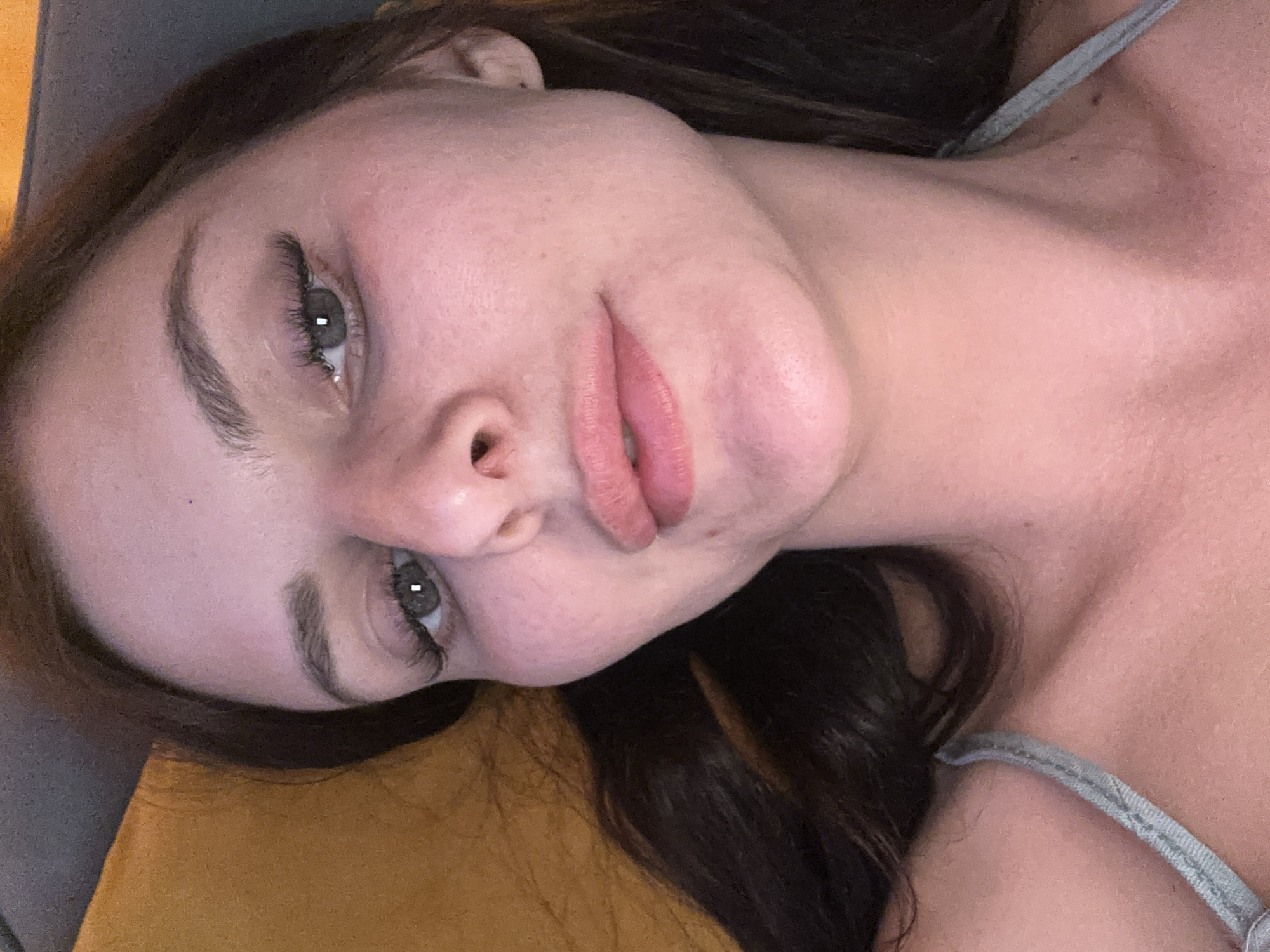 A close-up, frontal, chest-up, portrait-oriented, selfie-style photograph captures a young Caucasian woman lying down with her head tilted back and to the right. Her dark brown hair cascades to her left. She is wearing a light gray strap with a ribbed texture across her chest.
The woman's face is the central focus. Her eyes are open, looking towards the camera, with long, dark eyelashes. Her eyebrows are dark and well-defined, arched slightly. Her nose is prominent, and her lips are full and slightly parted, colored with a light pink lip gloss. Her skin appears fair with a scattering of freckles on her cheeks and a slight reddish hue on her cheeks and nose. Her neck and collarbone are visible. The background consists of a textured orange surface on the left and a dark gray surface on the top and right, suggestive of furniture or upholstery. The lighting appears to be dim, casting soft shadows. The overall impression is one of a casual, relaxed moment.