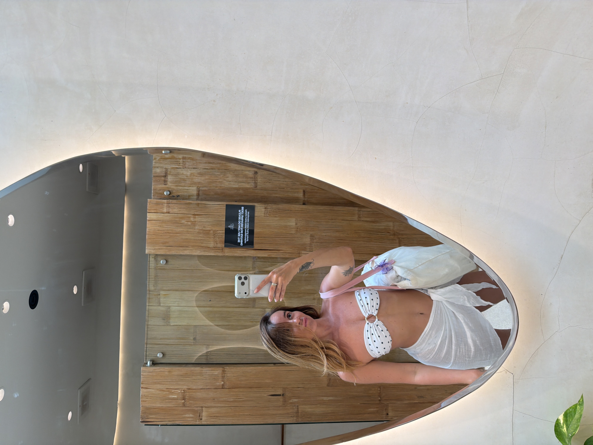 This media file shows a reflection of a woman in a mirror. She is wearing a white polka dot bikini and a white sarong. She is holding a smartphone and taking a selfie. The mirror is large and oval-shaped, with a light illuminating its edge. Behind her reflection, the mirror shows a wall with a bamboo texture. There is a black sticker on the bamboo wall with white text on it. The woman has blonde hair and appears to be relaxed and happy. The setting appears to be a bathroom or dressing area, possibly in a hotel or resort given the modern lighting and bamboo decor. The overall mood is casual and summery. The text on the sticker is upside down and reversed due to being a reflection, but appears to be in English and possibly Indonesian.