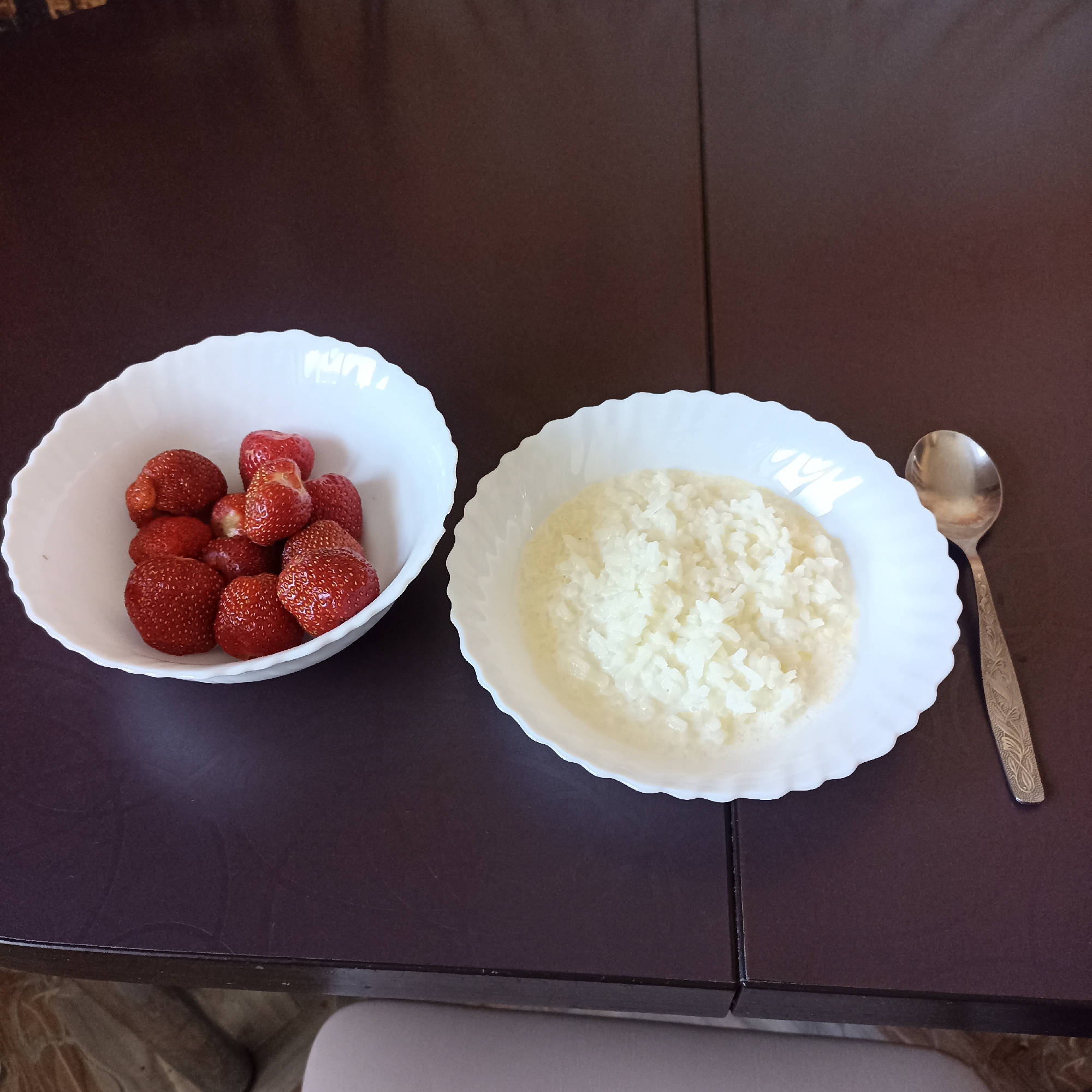 An overhead shot shows two white, fluted bowls and a spoon on a dark brown table. The bowl on the left contains about ten strawberries. The bowl on the right contains rice pudding. A spoon rests on the table to the right of the bowl of rice pudding. The table is dark brown and appears to be a wooden surface. The floor visible at the bottom of the image is tiled and a light gray chair is partially visible. The image is a photograph of breakfast food. This scene does not depict Shakhunya, Russia.