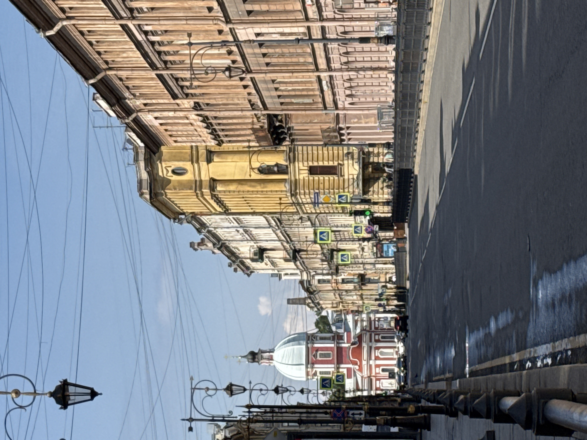 This is a vertical, medium-shot photograph of a street scene in Saint Petersburg, Russia. The perspective is from street level, looking down a wide asphalt road that curves slightly to the right. The road is lined with ornate lampposts on the left and a black metal fence on the right. The buildings lining the street are historic and display architectural details typical of European cities. On the left, a domed church with a red and white facade stands prominently. Several cars are visible on the road, and pedestrians can be seen walking on the sidewalks and near the buildings. The sky is clear and blue, with a few wisps of clouds. The overall impression is one of a sunny day in a historic urban setting.