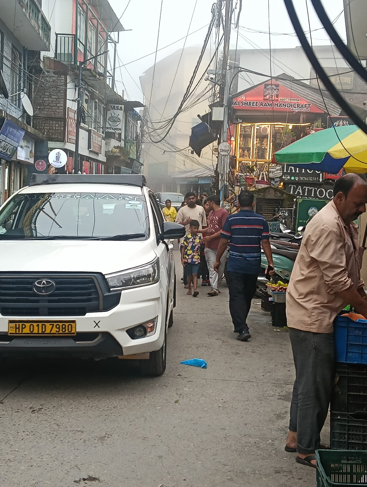 The image depicts a narrow, bustling street scene in Bhagsu, India, on a foggy day. A white Toyota Innova taxi, bearing the license plate "HP 01D 7980", is parked on the left side of the street. The taxi has "ARIHAAN" written on its windshield. People are walking along the street, some casually, while others appear to be shopping or working.
On the right side of the street, a man in a light brown shirt and dark pants is attending to stacked blue and green crates, some containing produce. In the background, shops with various signs are visible, including "DASA KITCHEN" and "AI SHREE RAM HANDICRAFT". The street is lined with multi-story buildings adorned with balconies and intricate wiring. The overall atmosphere is one of a vibrant market environment, possibly a tourist area given the handicraft and tattoo shop signs. The fog adds a soft, diffused quality to the light.