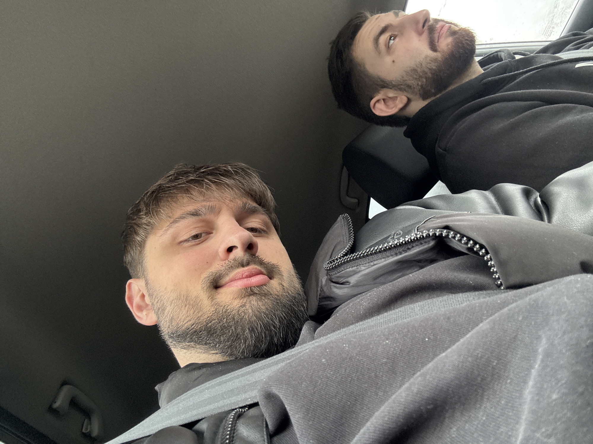 The image depicts the interior of a vehicle, occupied by two adult males. The specific location in the city of Bazan, Bulgaria, cannot be determined from the visual evidence provided.
The primary subject, positioned in the foreground and occupying the left side of the frame, is a male with light brown hair, a beard, and a mustache. He is wearing a black jacket, featuring a visible zipper, and a gray shoulder-belt strap is positioned across his chest. He faces the camera with a tilted head and his tongue slightly extended.
The secondary subject, located to the right and slightly behind the first, is a male with dark hair and a beard. He is wearing a black hooded sweatshirt and is observed in profile, looking towards the right side of the vehicle, out a window. A dark headrest is visible behind him.
The vehicle's interior features a dark headliner with a visible overhead grab handle. The exterior environment visible through the window next to the second subject is indistinct, appearing as a light gray, likely overcast, sky or distant scene.