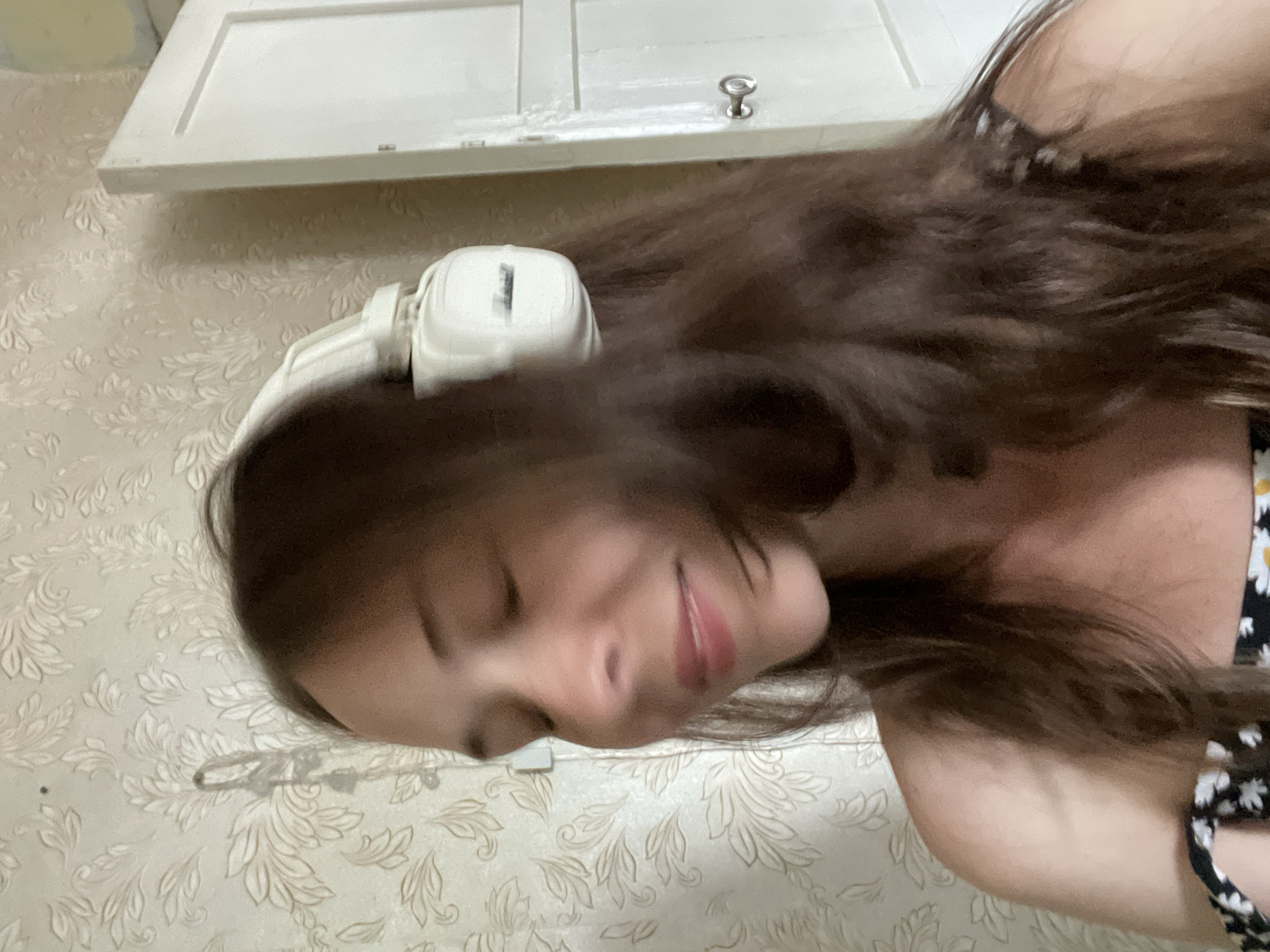 A medium shot shows a young woman with long brown hair and closed eyes wearing white headphones. She is positioned in the foreground, with her head turned to the right, smiling slightly. Her shoulders are visible, and she is wearing a black and white floral patterned top. In the background, a wall with floral patterned wallpaper is visible, and to the right, a white door with a silver doorknob. The lighting appears to be natural, coming from the front and right. The image is slightly blurred due to motion. The scene is indoors, in what appears to be a residential setting.