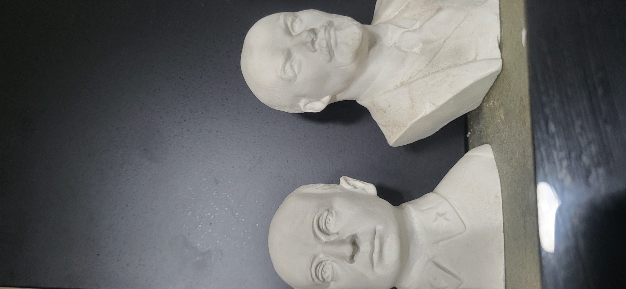Two white plaster busts lie on a dark surface. The bust on the right is partially cropped off at the top, while the one on the bottom is more fully visible. The bottom bust has what appears to be a star on the lapel. The background is a dark, mottled gray. The image contains no specific location clues that would indicate Moscow, Russia.
