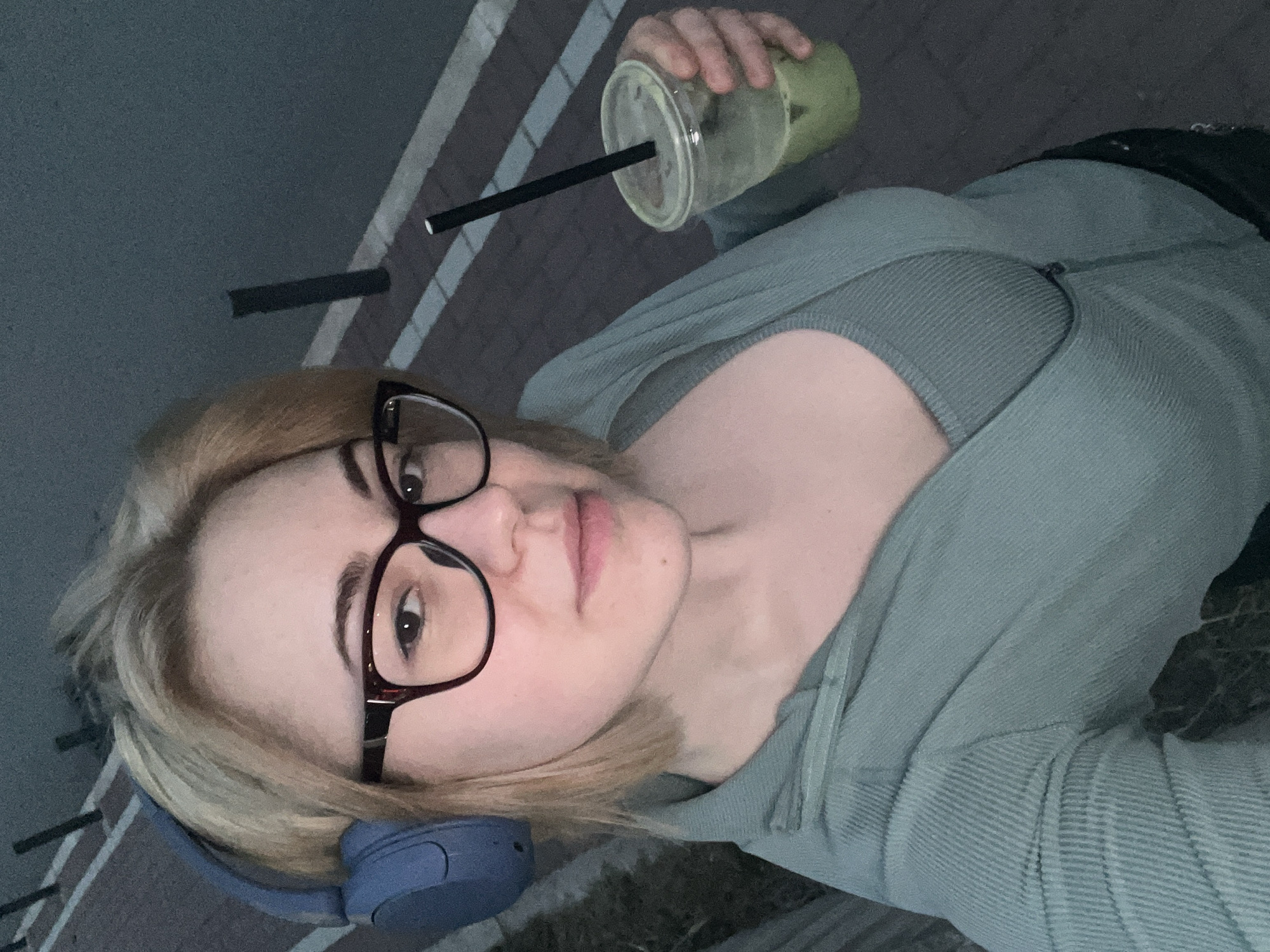 A woman is outdoors holding a cup with a straw. She is wearing glasses, headphones, and a green hoodie. The location is Boreti, Montenegro.