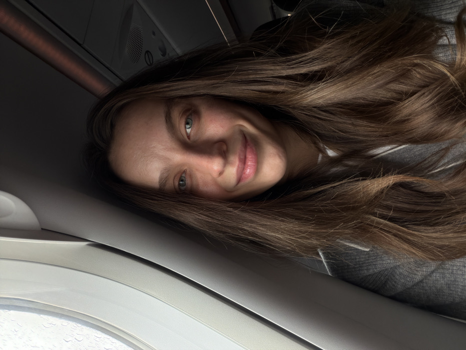 This image depicts a young adult female inside an aircraft cabin, located in Mojanovići, Montenegro. The female subject has long, wavy brown hair with lighter highlights, parted centrally. She is smiling directly at the camera with closed lips, showing dimples on both cheeks. Her light-colored eyes are visible, and her complexion is fair, with a slight flush on her nose and cheeks. She wears a grey ribbed garment, possibly a sweater or hoodie, with a white drawstring visible near the neckline. To the left, an aircraft window displays water droplets on its exterior surface against an indistinct bright background. The upper right portion of the scene reveals dark overhead paneling consistent with an aircraft interior, with a horizontal strip of dim reddish-orange light visible.