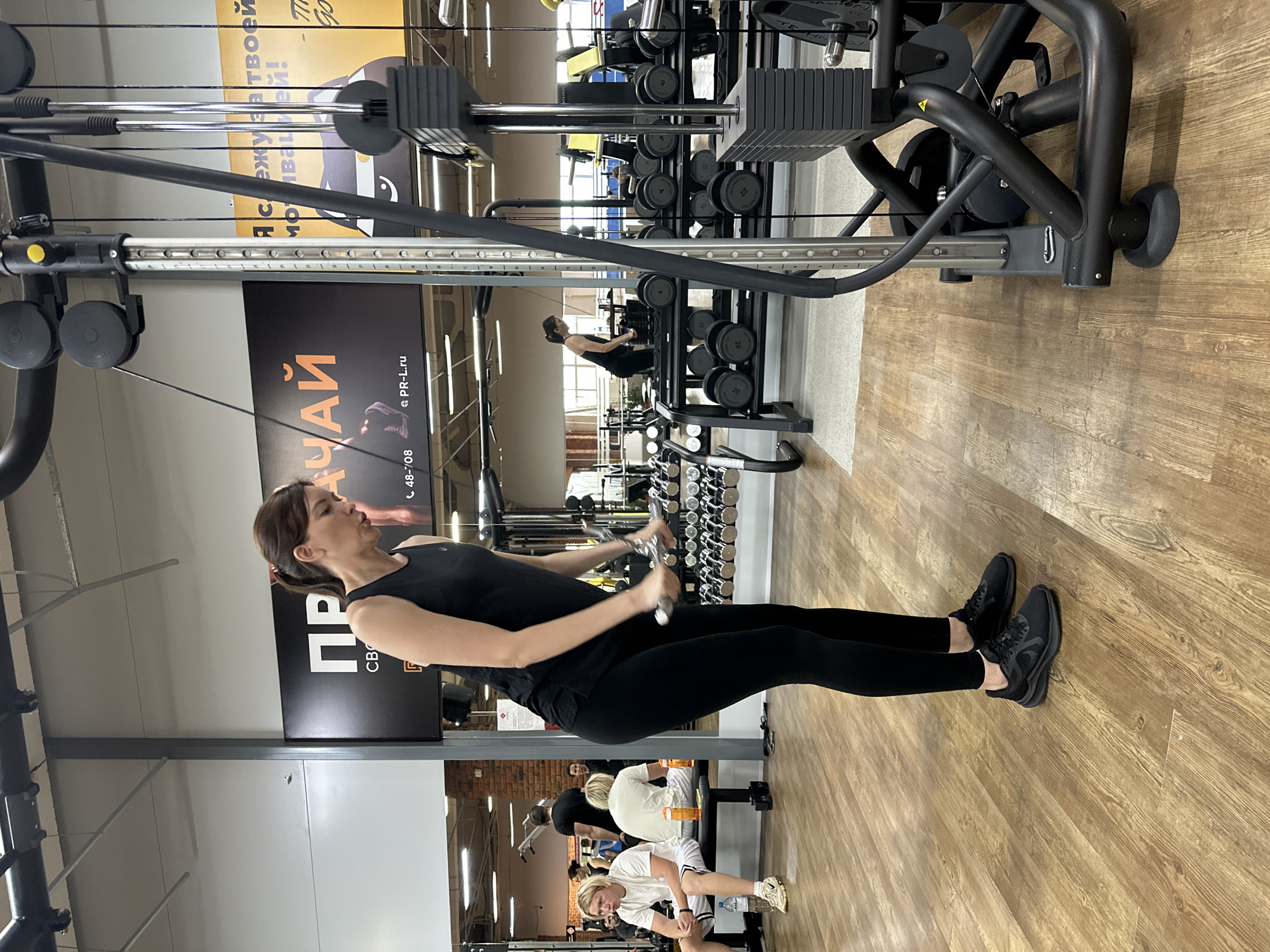 The image shows a woman in a gym performing an exercise with a cable machine. She is wearing a black tank top and black leggings, with black sneakers. The machine has weight plates and a handle she is holding. Behind her is a rack of dumbbells. Other people are present in the gym, some sitting on benches. The location is likely a gym in Chaikovsky, Russia, based on the Cyrillic text "ЧАЙ" visible in the image and the domain "PR-L.ru".