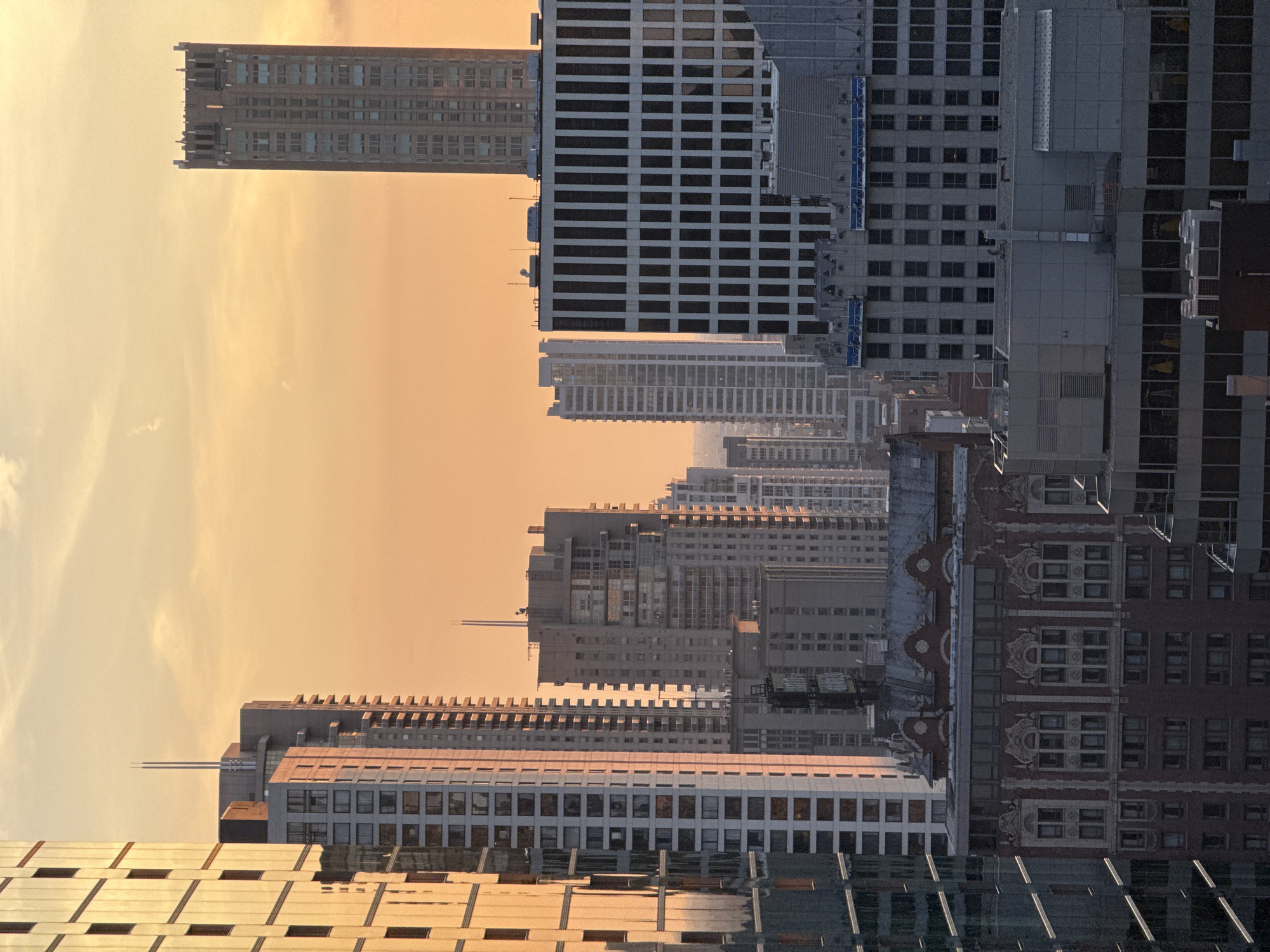 The image depicts a cityscape at sunset, with a warm orange glow illuminating the sky and reflecting off the numerous skyscrapers. The buildings are a mix of modern glass and steel structures and older brick buildings. In the foreground, a tall, modern building with a glass facade dominates the left side of the frame. To its right, several other high-rise buildings of varying heights and architectural styles are visible. The most prominent structure on the right side is a distinctive tower with a decorative crown. The scene is devoid of people, and the overall impression is one of urban grandeur and the quiet beauty of a city at dusk.
