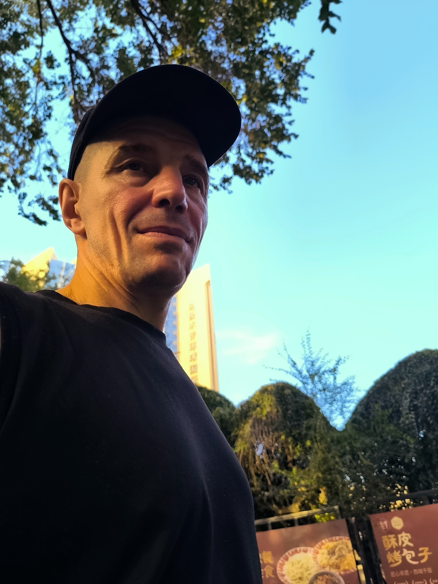 An outdoor, eye-level, medium shot shows a bald man wearing a black baseball cap and a black t-shirt. He is looking up and to the right. The man is illuminated by golden hour lighting, casting shadows on his face. In the background, there are trees, a tall, light-colored building, and a blue sky. To the right of the man, there are two dark green, rounded bushes and a sign with Chinese characters and images of food. The image appears to have been taken in Ürümqi, China, based on the presence of Chinese text on the sign.