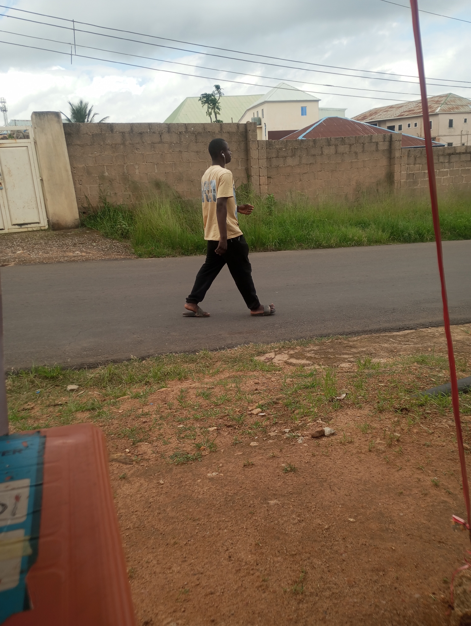 A young Black male wearing a tan t-shirt and black pants walks on a paved road with a red pole on the right side of the frame. He is wearing sandals on his feet. The background consists of a brown brick wall, tall green grass, and several buildings with corrugated metal roofs, indicating an outdoor urban setting. The sky is overcast. The image is taken from a low angle, with some of the foreground obscured by a reddish-brown surface, possibly a bench or table. The overall scene suggests a residential or commercial area in Jos, Nigeria.