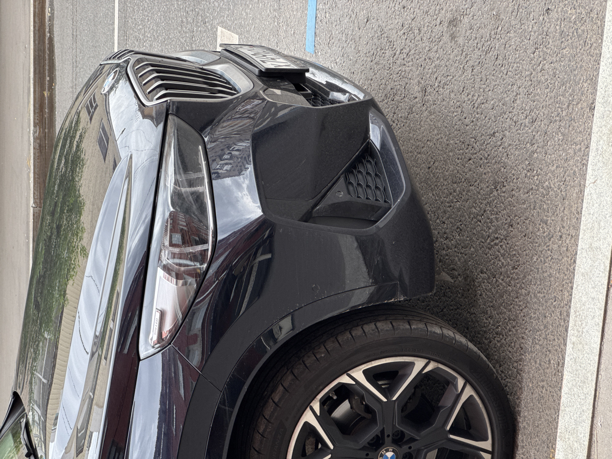 A close-up, eye-level, outdoor shot shows the front right quarter of a dark-colored BMW SUV parked on a paved street in Moscow, Russia. The car's grille, headlight, and front wheel are prominent. The pavement is a grey asphalt with white and blue markings indicating a parking space. Reflections of buildings are visible on the car's hood and body. The car appears to be in good condition, with no visible damage. The environment is urban, with a sense of a city street on a clear day.