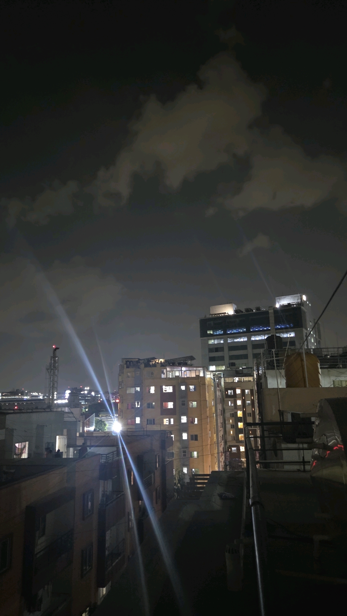 An urban cityscape at night, likely in Bengaluru, India, showcases buildings of varying heights illuminated by interior lights and streetlights. The sky is dark with scattered clouds, some of which are catching the light. Two prominent beams of light, possibly from searchlights or strong spotlights, cut across the scene from the lower left towards the upper right. In the foreground, a rooftop is visible with pipes and cables. Several buildings are discernible, including a tall, modern building with many lit windows in the mid-right, and a cluster of residential buildings with numerous windows, some lit with warm light and others dark, to the left of the modern building. A metallic tower with a red light at its apex is visible in the far left distance. The overall impression is one of urban nocturnal activity and architectural diversity.