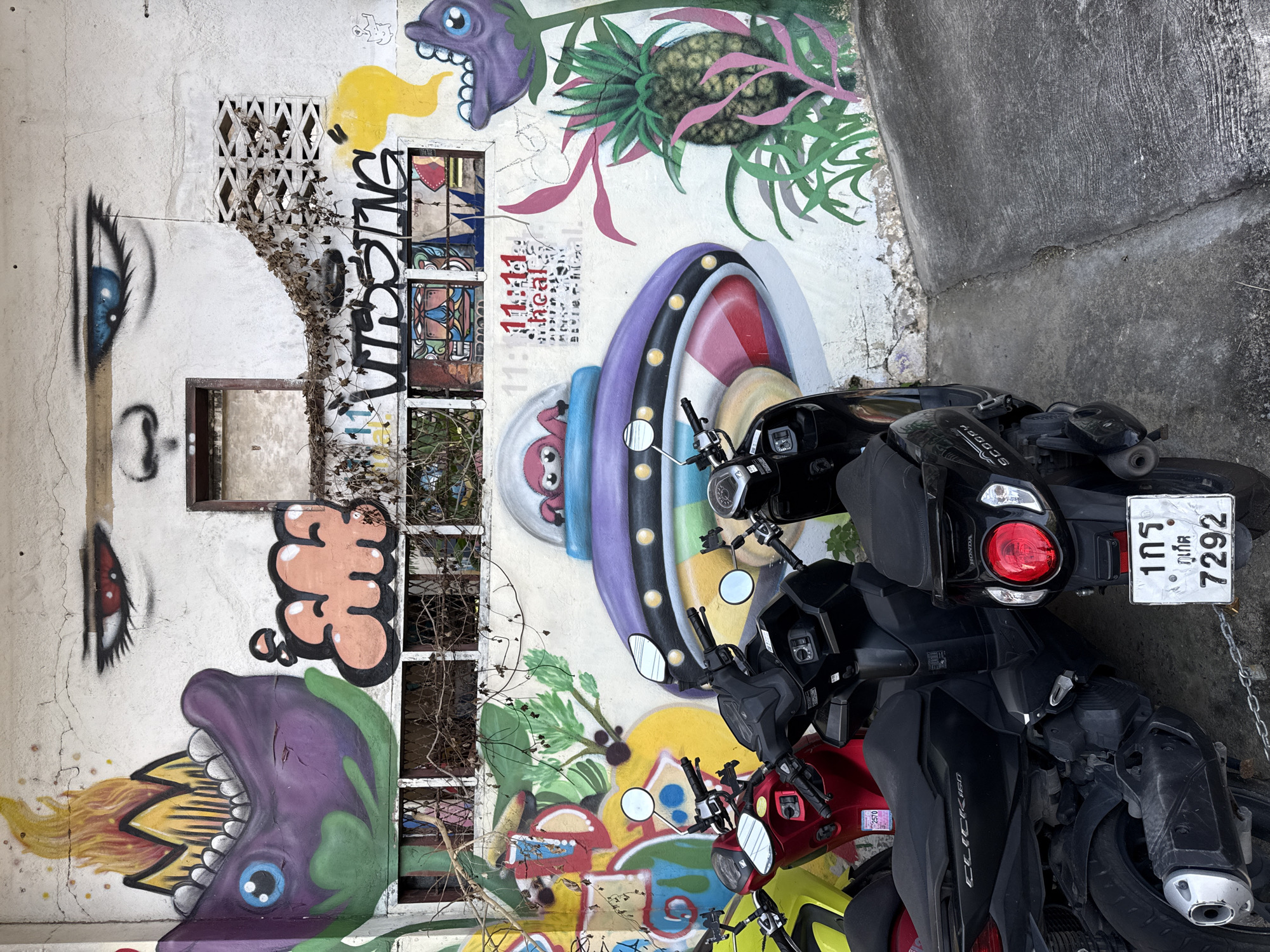 The image shows a section of a white, weathered wall covered in graffiti and artwork. The graffiti includes cartoonish faces, a large eye with blue iris, flames, a purple alien-like creature with fangs, and a UFO with an alien inside. There are also images of plants, including a pineapple, and text that appears to be "MISSING" and "11 11:11 heal." Several motorcycles are parked in front of the wall, obscuring the lower portion of the artwork and the wall's base. The ground in front of the wall is concrete, with cracks and some debris. The scene appears to be an outdoor urban environment, likely in a back alley or a less maintained area. The license plate on the black motorcycle in the foreground is visible, with Thai characters and the number 7292. Based on the style of the graffiti and the license plate, the location is likely within the city of Phuket City Municipality, Thailand.