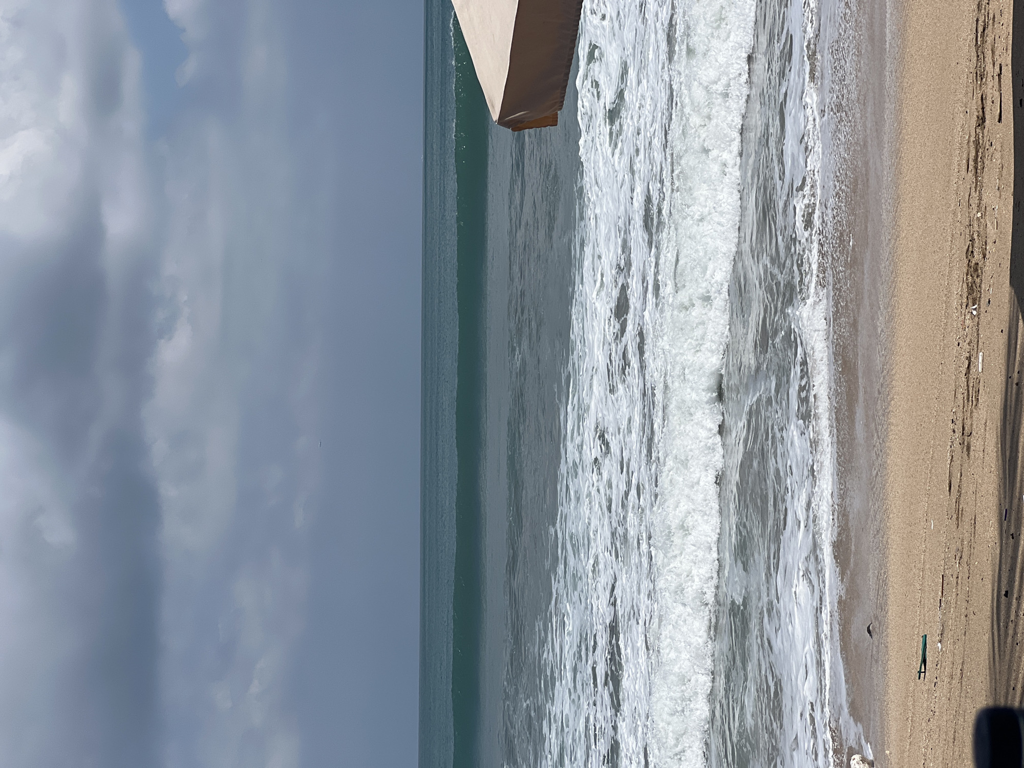 An overcast sky looms over a tranquil ocean scene in Kuta, Indonesia. The vast expanse of the sea is characterized by a series of waves, ranging from a gentle swell further out to frothy, white-capped breakers closer to the shore. The water transitions from a deep, dark teal in the distance to a lighter, more agitated green and white near the beach.
The foreground is dominated by a sandy beach, speckled with small debris and shadows cast by an unseen object, possibly a beach umbrella, partially visible in the upper right corner. The umbrella is a pale, sandy color and appears to be made of fabric. The sand itself is a light tan, with areas of dampness where the waves have receded. The overall atmosphere suggests a calm, perhaps slightly cool day, with the clouds obscuring direct sunlight. There are no people visible in the frame, and the scene is devoid of any discernible activity beyond the natural movement of the waves.